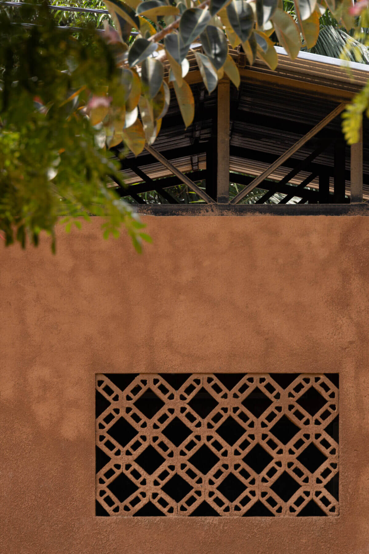 Exterior da escola com parede texturizada em tom de terra e janela gradeada, sob teto metálico e folhagem. — Escola Kapalanga arquitetura Paulo Moreira Architectures e PARQ Arquitectos em Luanda