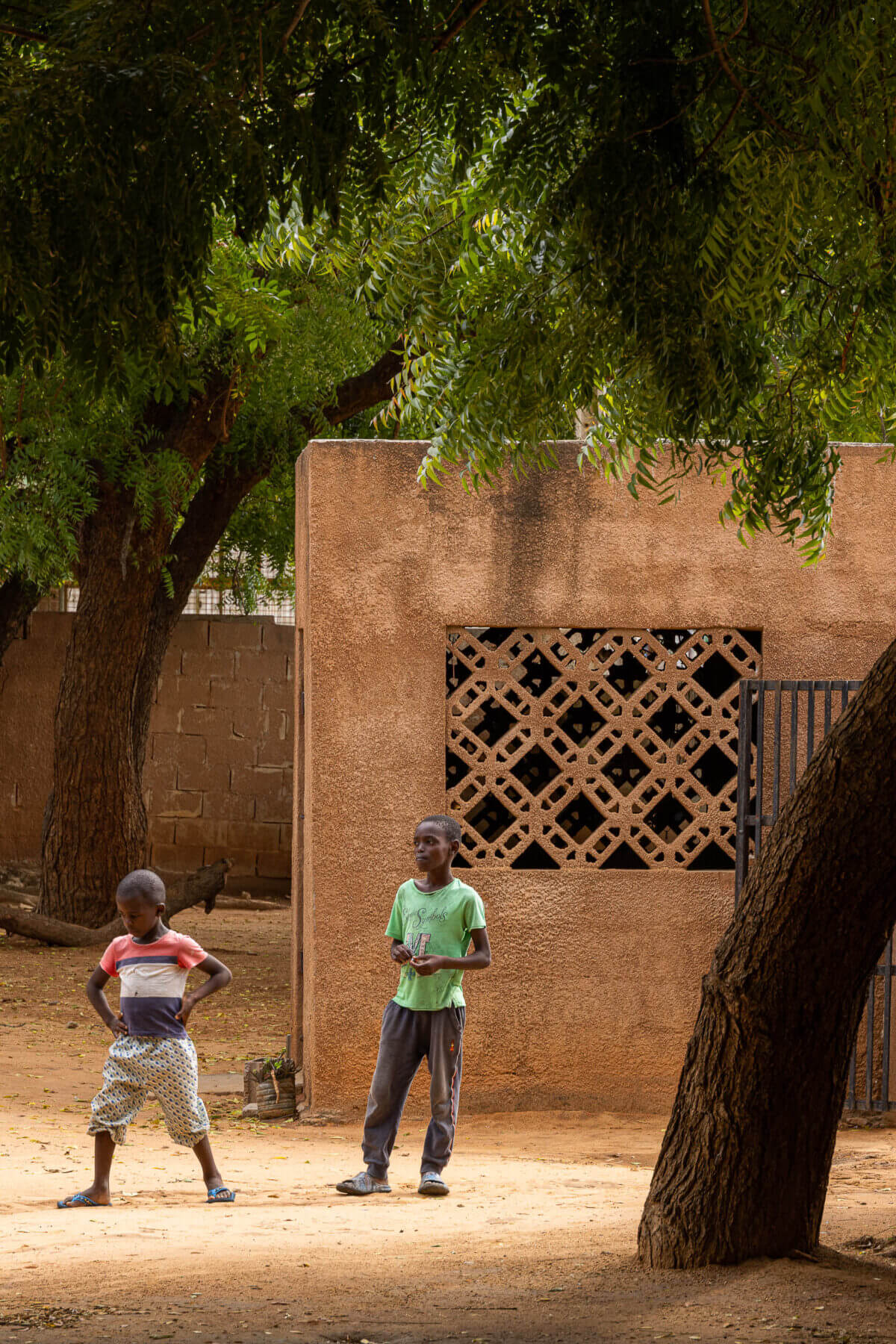 Exterior da escola com crianças em frente a um edifício de paredes texturizadas em tom de terra e janela gradeada. — Escola Kapalanga arquitetura Paulo Moreira Architectures e PARQ Arquitectos em Luanda