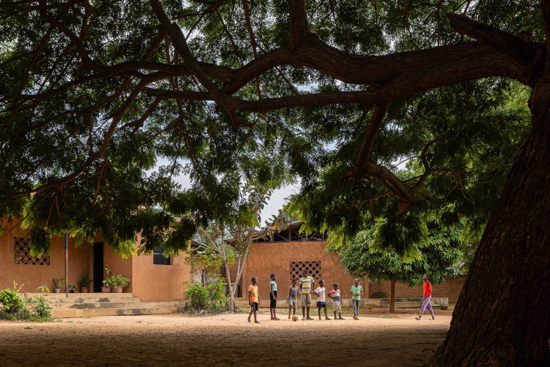 Pátio da escola com crianças a jogar futebol sob a sombra de uma grande árvore, com edifícios escolares ao fundo. — Escola Kapalanga arquitetura Paulo Moreira Architectures e PARQ Arquitectos em Luanda