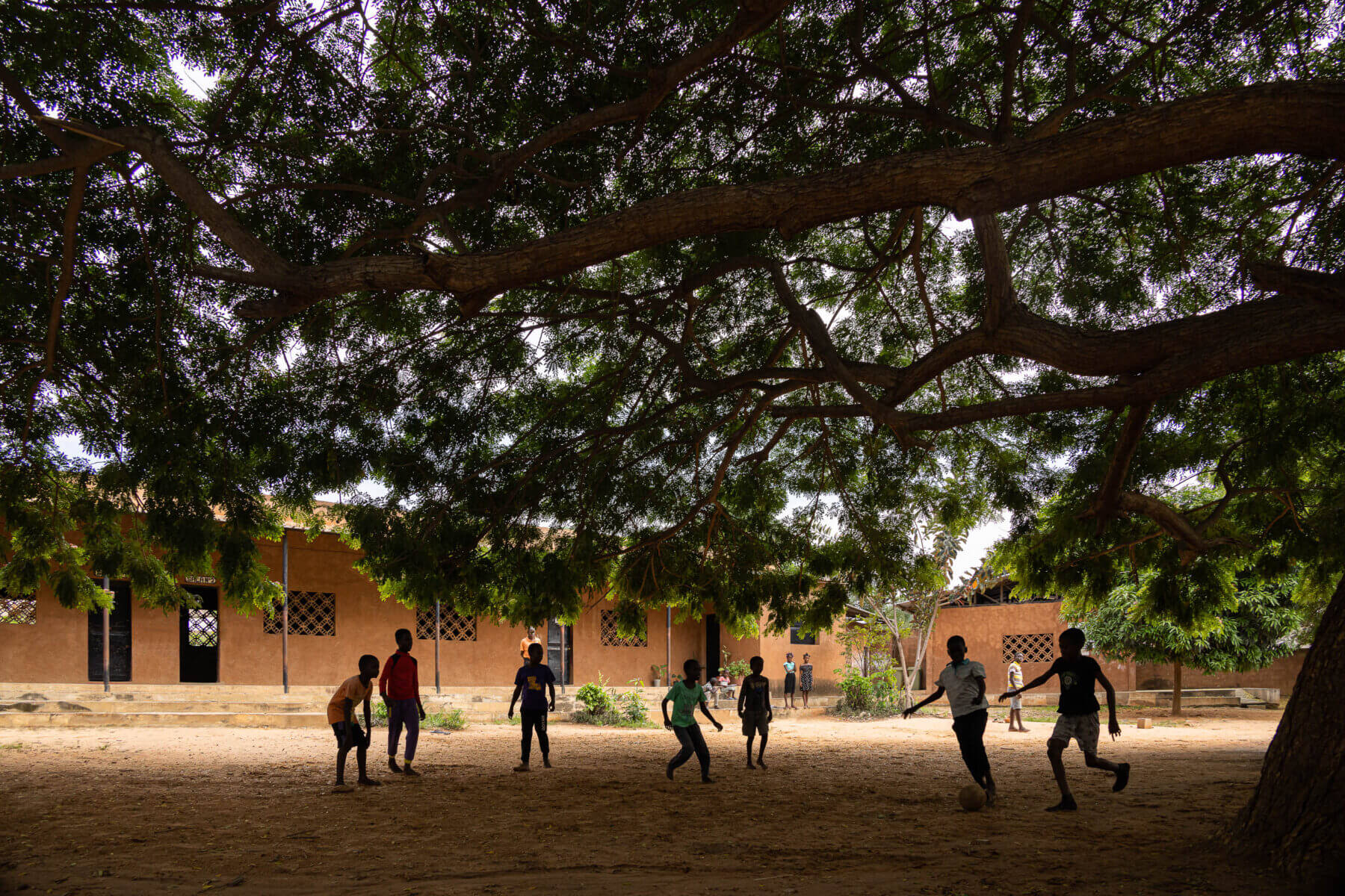 Pátio da escola com crianças a jogar futebol sob a sombra de uma grande árvore, com edifícios escolares ao fundo. — Escola Kapalanga arquitetura Paulo Moreira Architectures e PARQ Arquitectos em Luanda