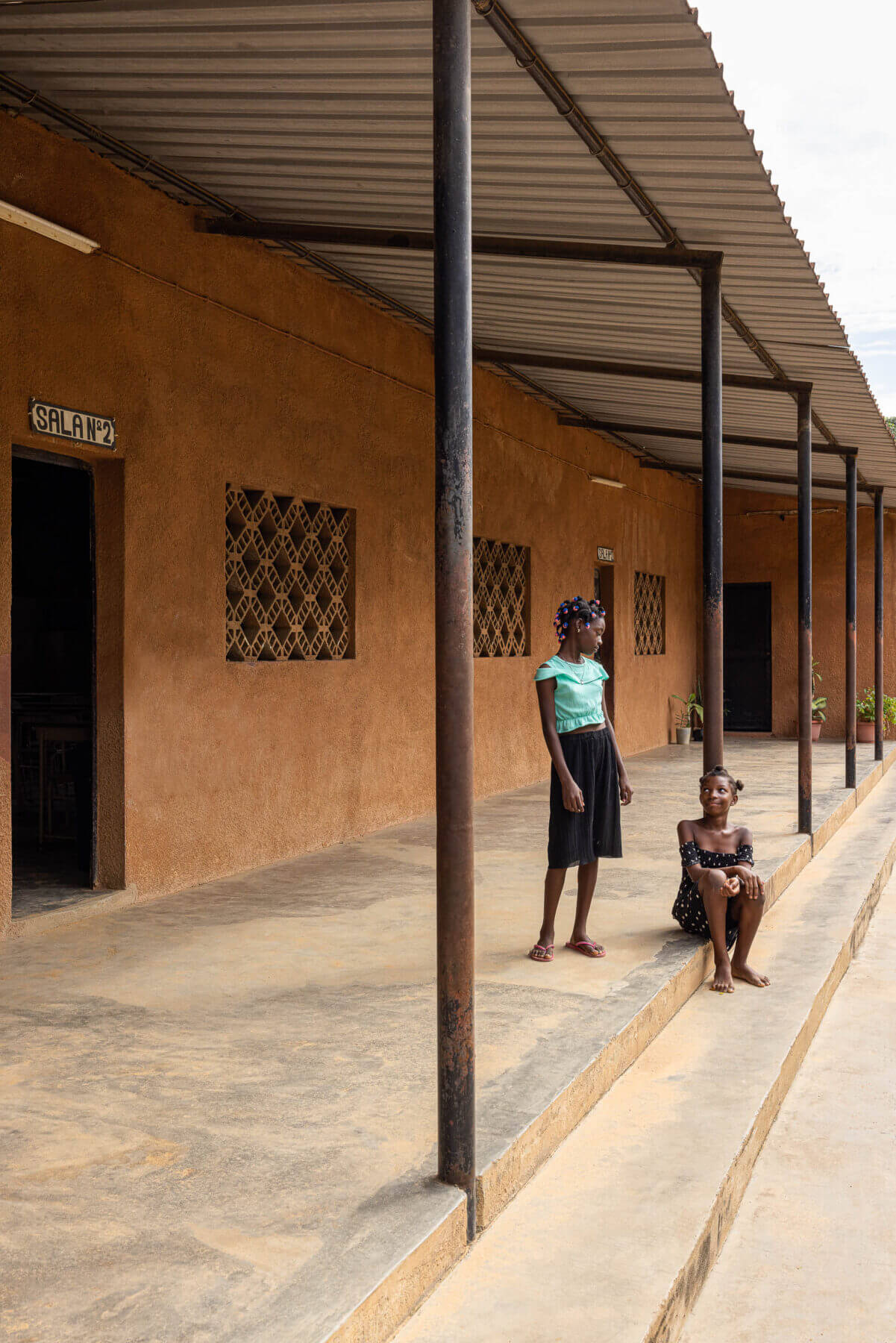 Alpendre com pilares metálicos, paredes texturizadas em tom de terra e janelas gradeadas, com crianças no espaço. — Escola Kapalanga arquitetura Paulo Moreira Architectures e PARQ Arquitectos em Luanda