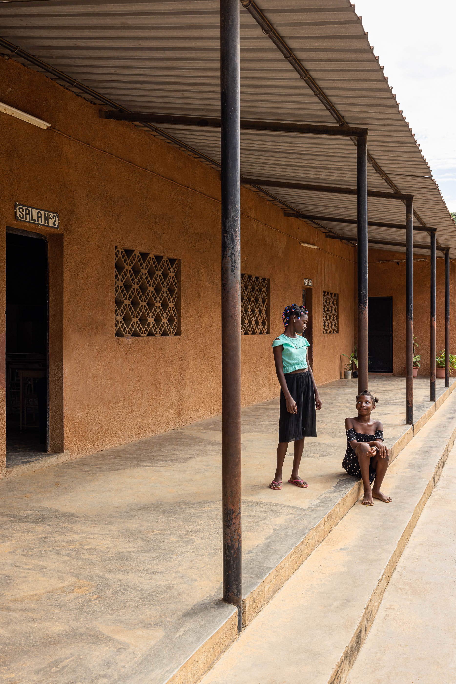 Alpendre com pilares metálicos, paredes texturizadas em tom de terra e janelas gradeadas, com crianças no espaço. — Escola Kapalanga arquitetura Paulo Moreira Architectures e PARQ Arquitectos em Luanda