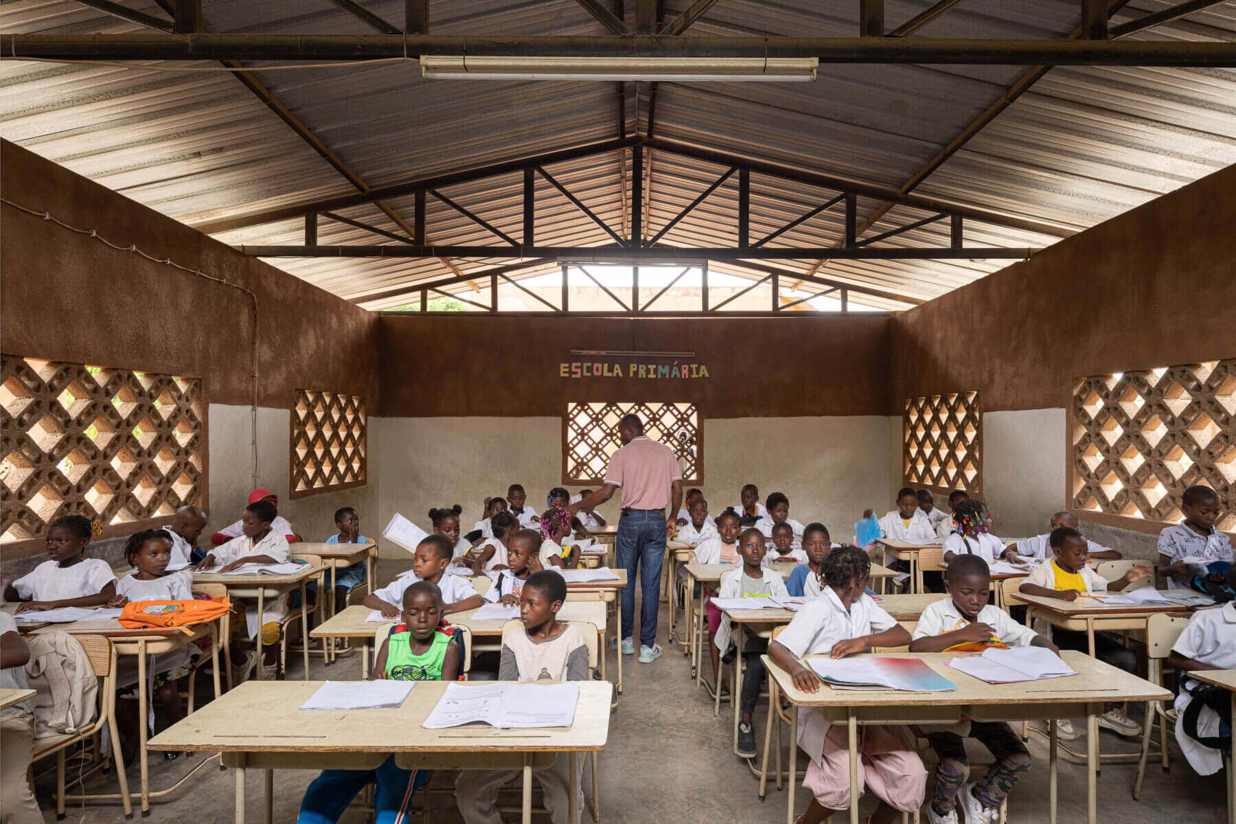 Sala de aula cheia de alunos e um professor, com carteiras de madeira, janelas gradeadas e paredes em dois tons. — Escola Kapalanga arquitetura Paulo Moreira Architectures e PARQ Arquitectos em Luanda