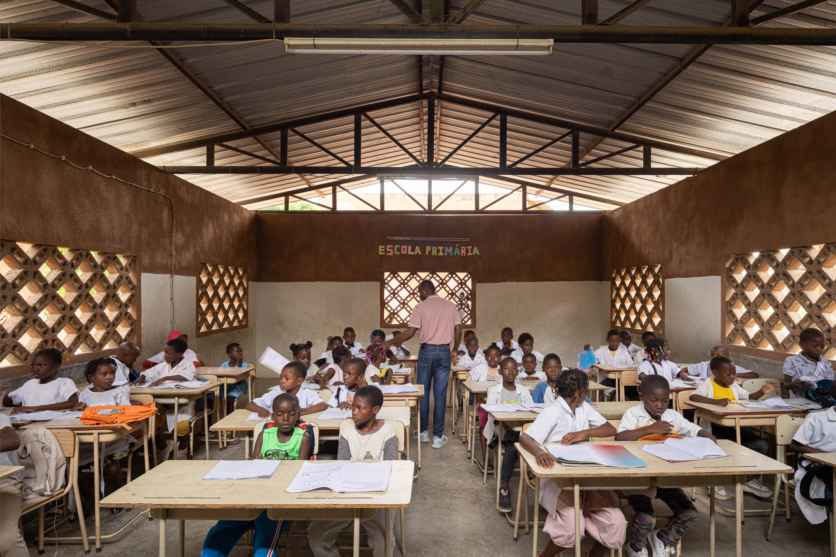 Sala de aula cheia de alunos e um professor, com carteiras de madeira, janelas gradeadas e paredes em dois tons. — Escola Kapalanga arquitetura Paulo Moreira Architectures e PARQ Arquitectos em Luanda