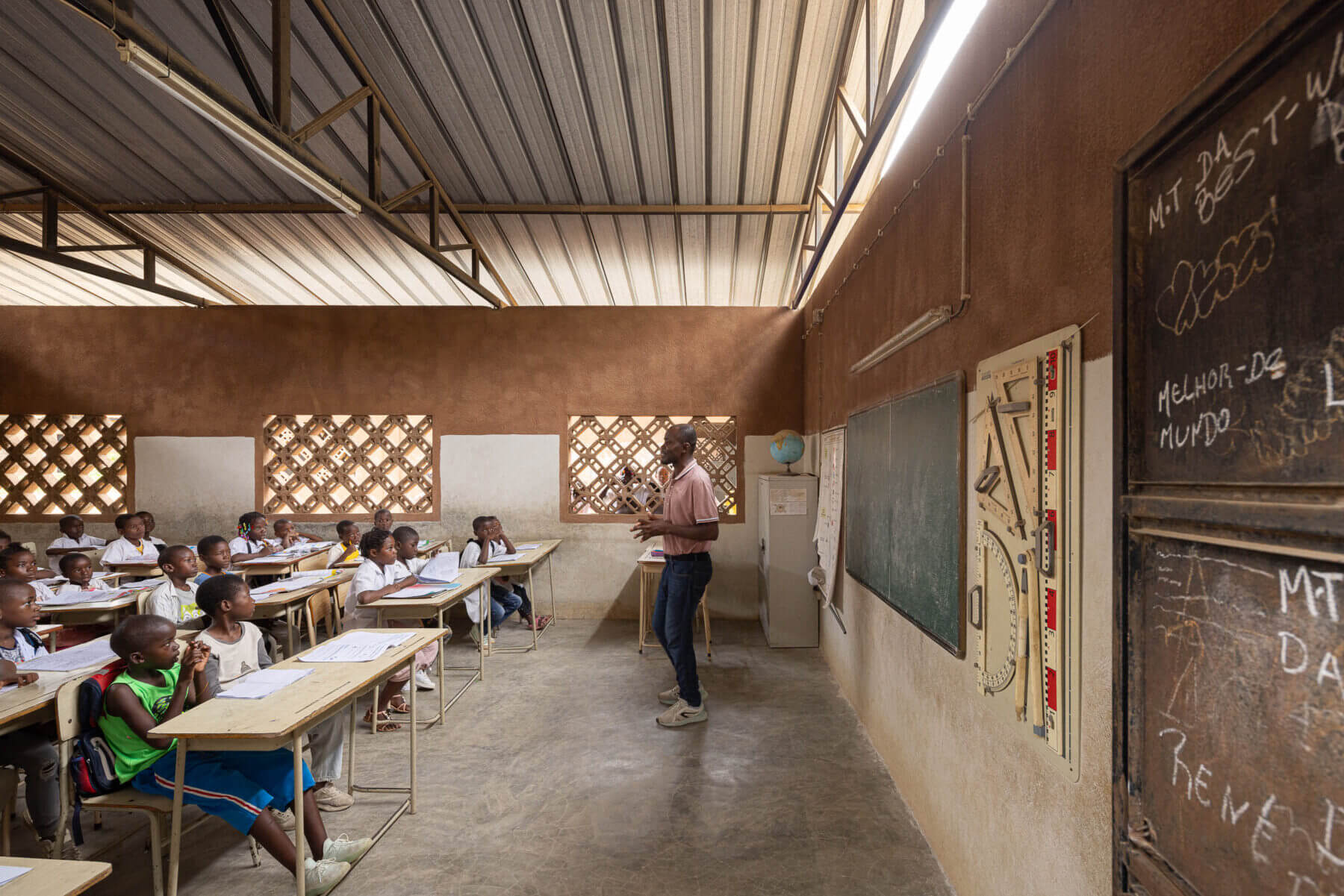 Sala de aula com alunos e um professor, com quadro negro, material didático e janelas gradeadas. — Escola Kapalanga arquitetura Paulo Moreira Architectures e PARQ Arquitectos em Luanda