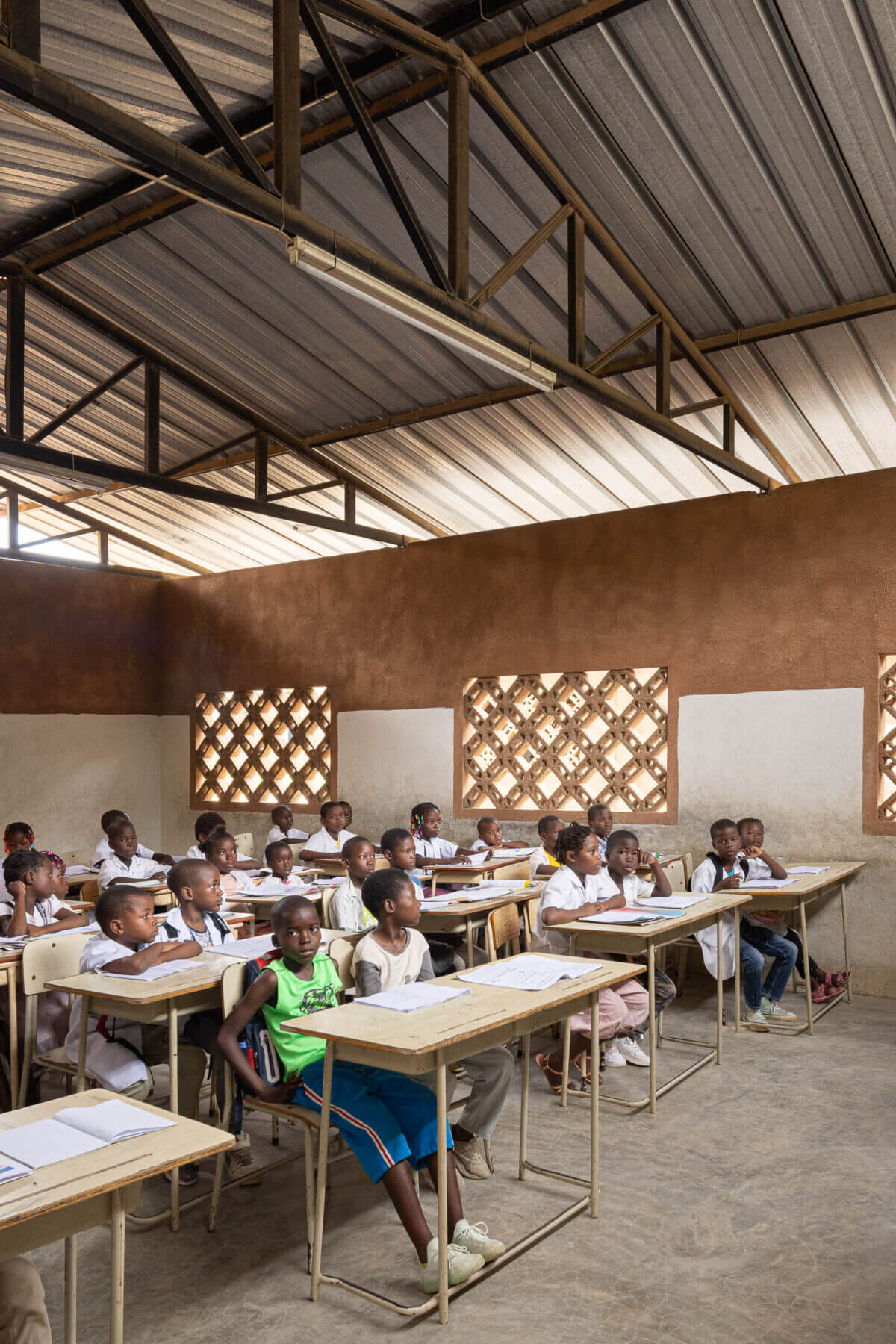 Sala de aula com alunos sentados nas carteiras de madeira, janelas gradeadas e paredes em dois tons. — Escola Kapalanga arquitetura Paulo Moreira Architectures e PARQ Arquitectos em Luanda