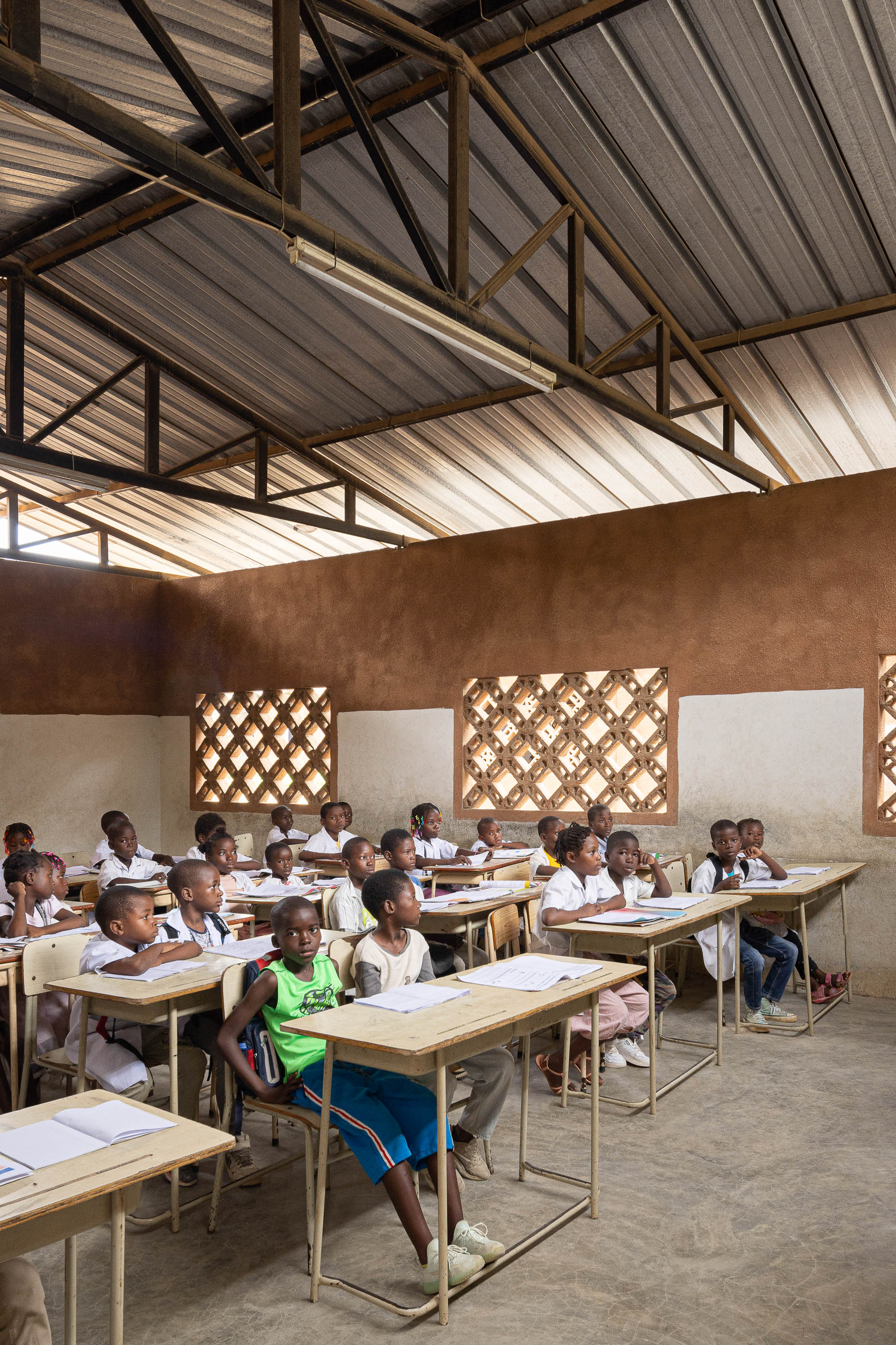 Sala de aula com alunos sentados nas carteiras de madeira, janelas gradeadas e paredes em dois tons. — Escola Kapalanga arquitetura Paulo Moreira Architectures e PARQ Arquitectos em Luanda