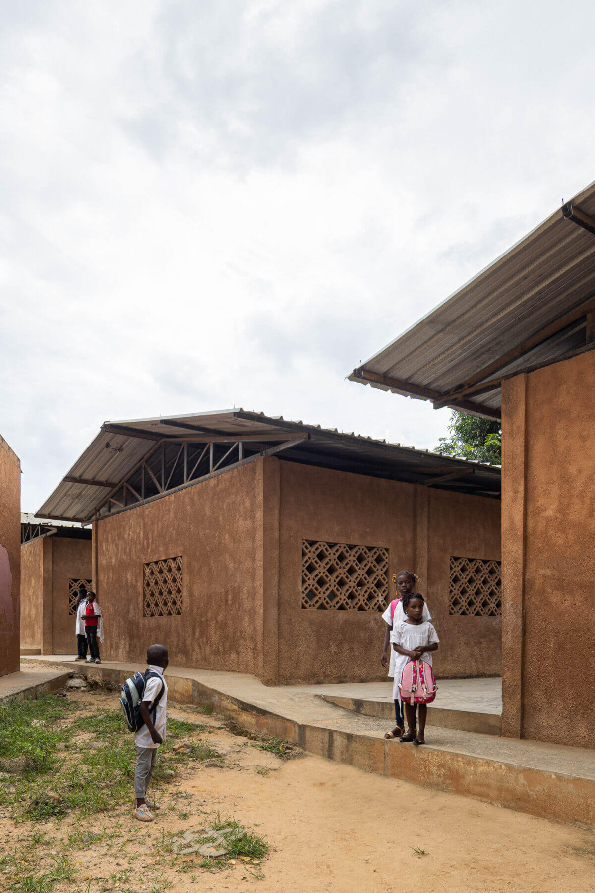 Exterior da escola com edifícios térreos de paredes texturizadas em tons de terra e janelas gradeadas, com crianças. — Escola Kapalanga arquitetura Paulo Moreira Architectures e PARQ Arquitectos em Luanda