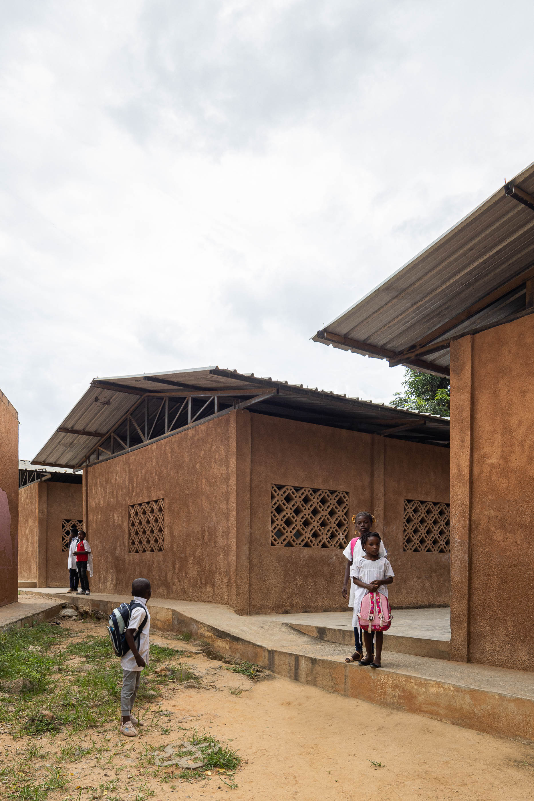 Exterior da escola com edifícios térreos de paredes texturizadas em tons de terra e janelas gradeadas, com crianças. — Escola Kapalanga arquitetura Paulo Moreira Architectures e PARQ Arquitectos em Luanda