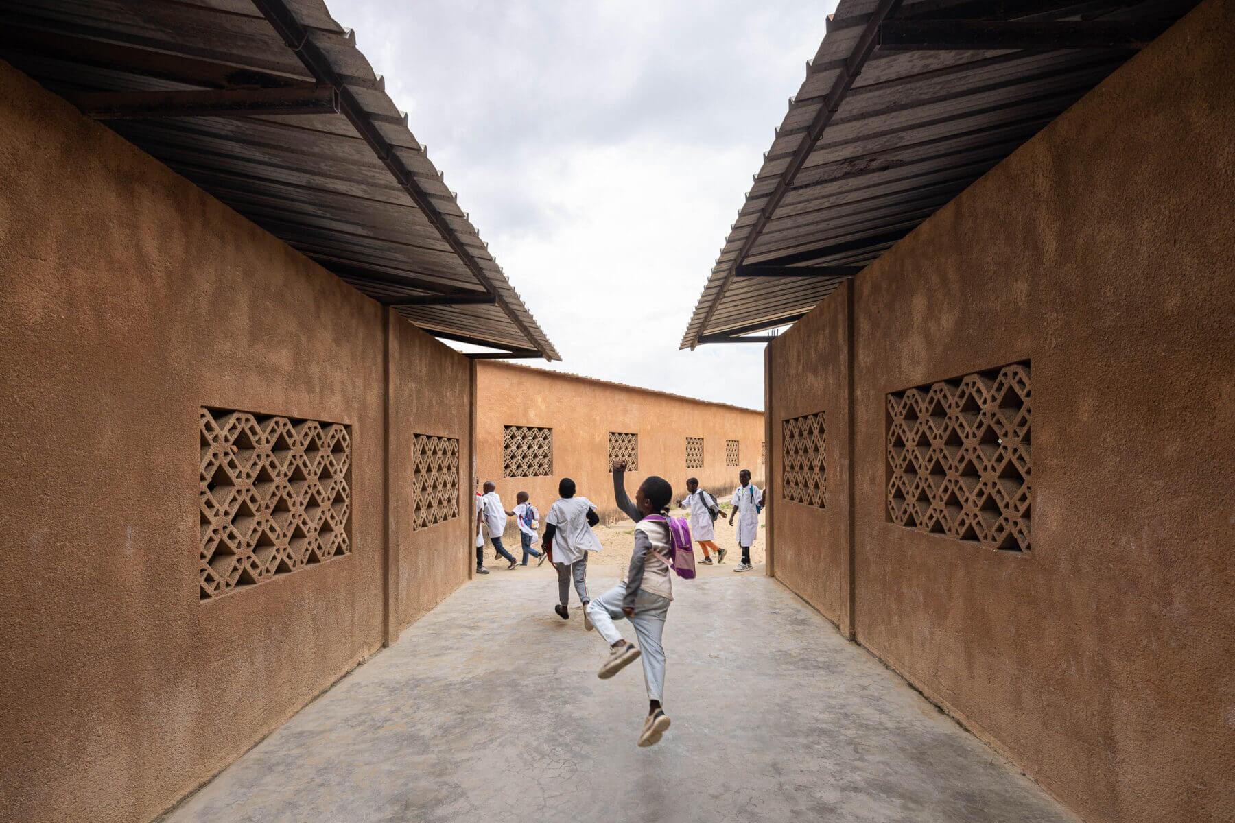 Corredor exterior entre edifícios escolares com paredes texturizadas em tom de terra e janelas gradeadas, com crianças. — Escola Kapalanga arquitetura Paulo Moreira Architectures e PARQ Arquitectos em Luanda