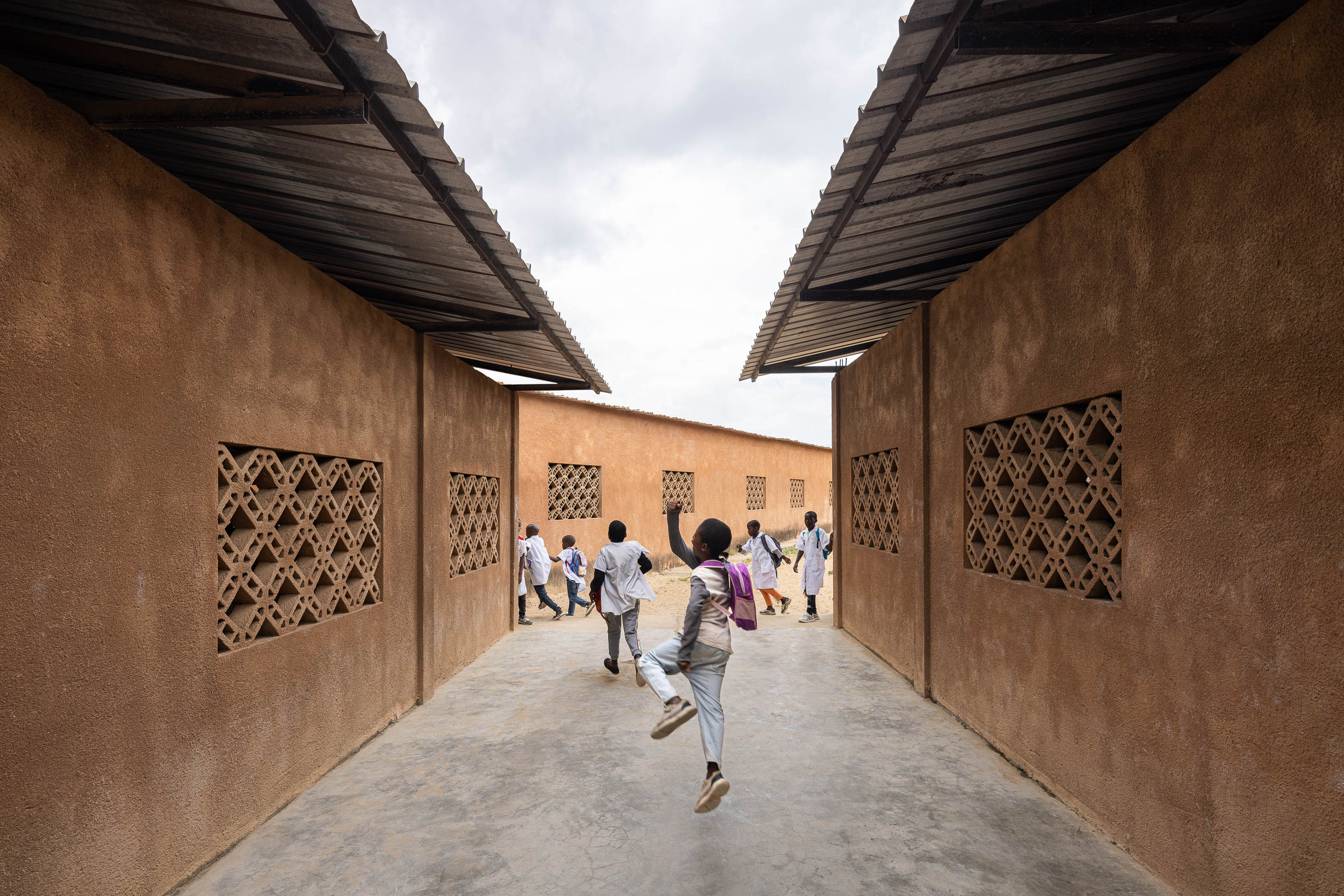 Corredor exterior entre edifícios escolares com paredes texturizadas em tom de terra e janelas gradeadas, com crianças. — Escola Kapalanga arquitetura Paulo Moreira Architectures e PARQ Arquitectos em Luanda