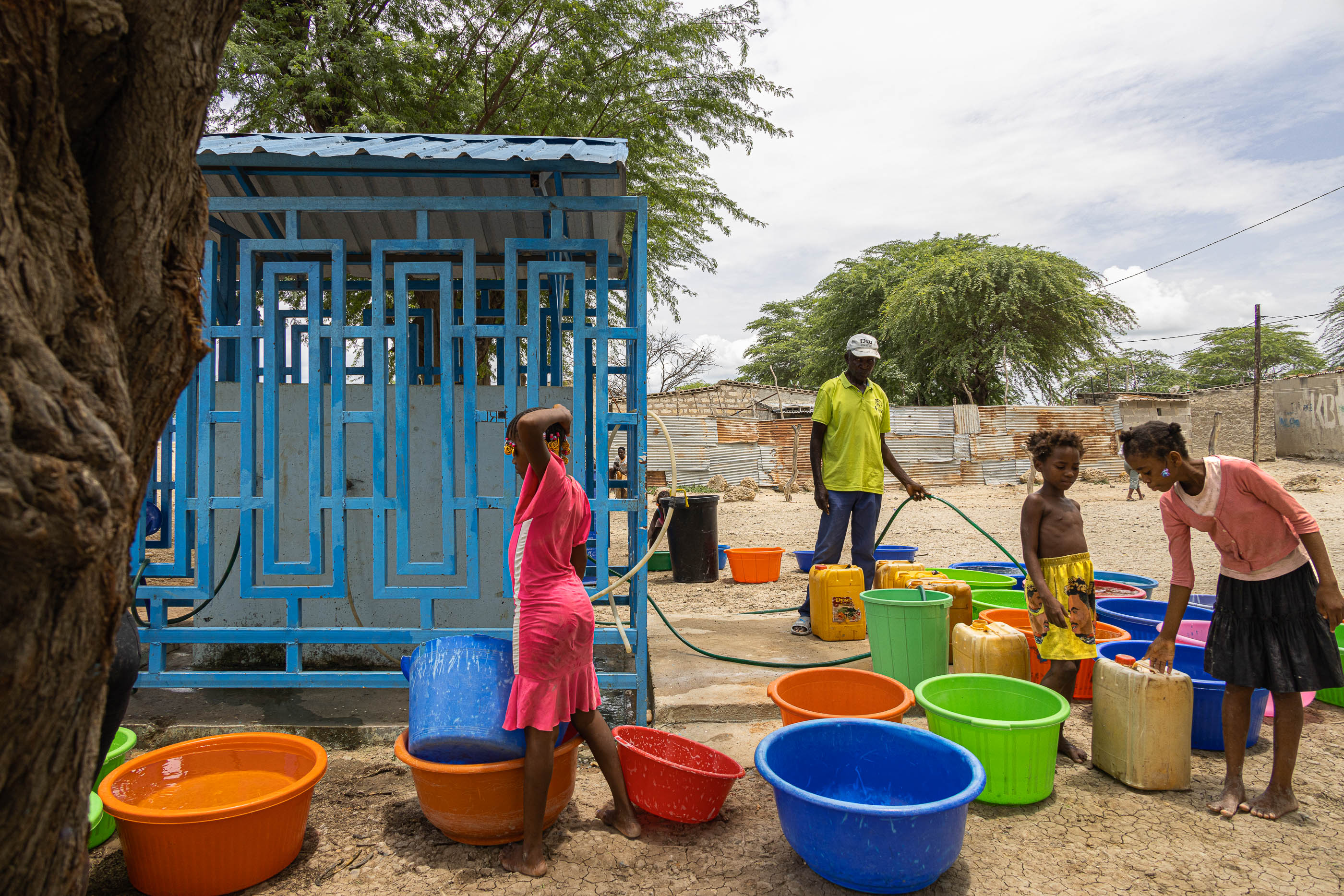 Chafariz: Pessoas a recolher água num chafariz azul, com muitos baldes coloridos no chão. — Chafarizes em Luanda arquitetura Paulo Moreira Architectures em Luanda