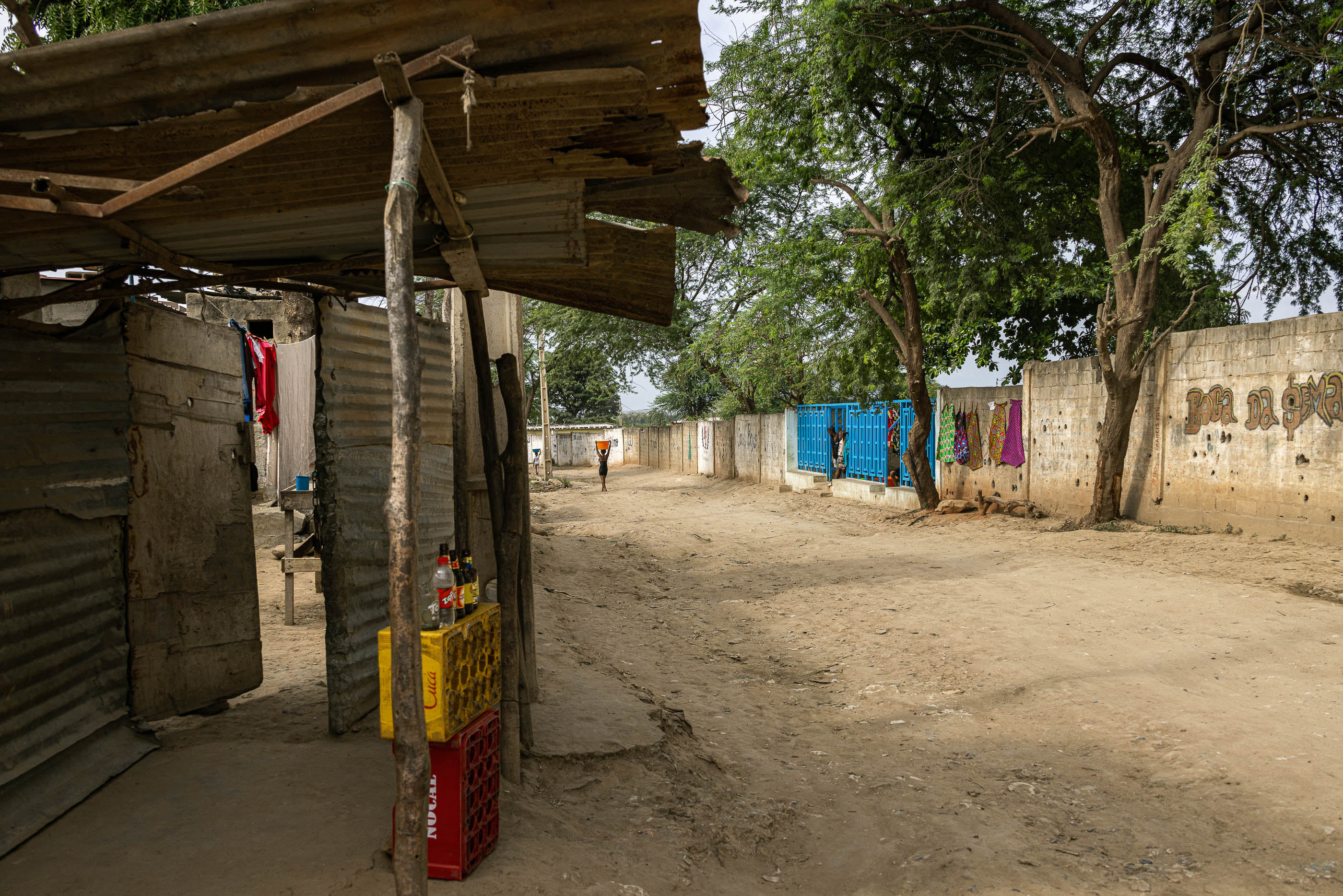 Exterior: Rua de terra batida com um chafariz ao fundo, barracas improvisadas e roupa a secar. — Chafarizes em Luanda arquitetura Paulo Moreira Architectures em Luanda