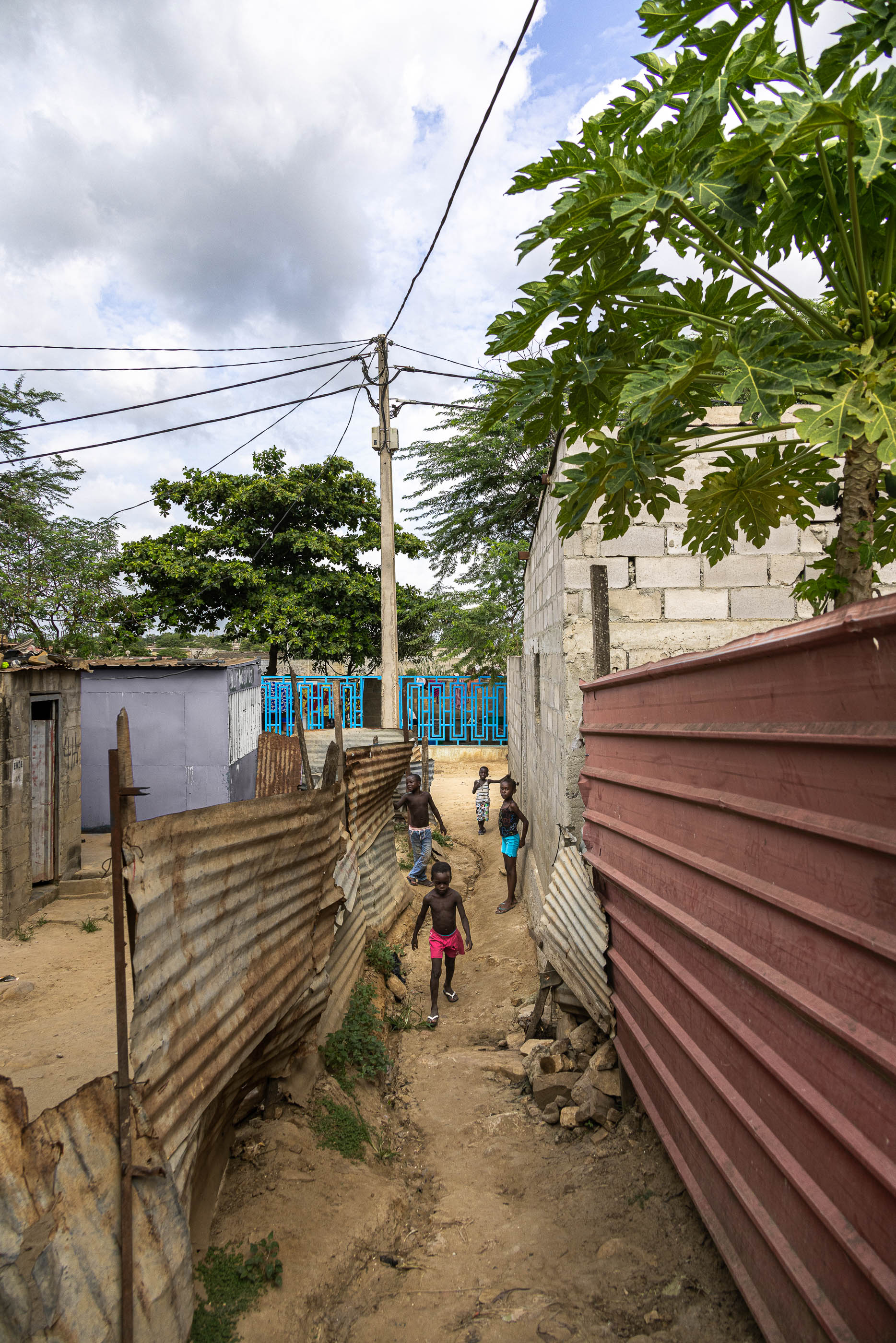 Rua: Beco estreito entre casas de zinco e blocos de cimento, com crianças a passar. — Chafarizes em Luanda arquitetura Paulo Moreira Architectures em Luanda