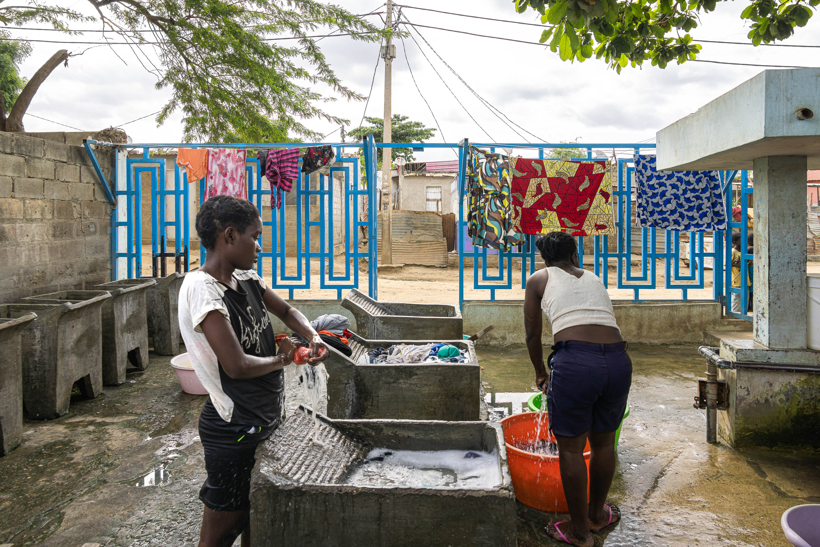 Lavandaria: Mulheres a lavar roupa em tanques de cimento, com roupa a secar e pavimento molhado. — Chafarizes em Luanda arquitetura Paulo Moreira Architectures em Luanda