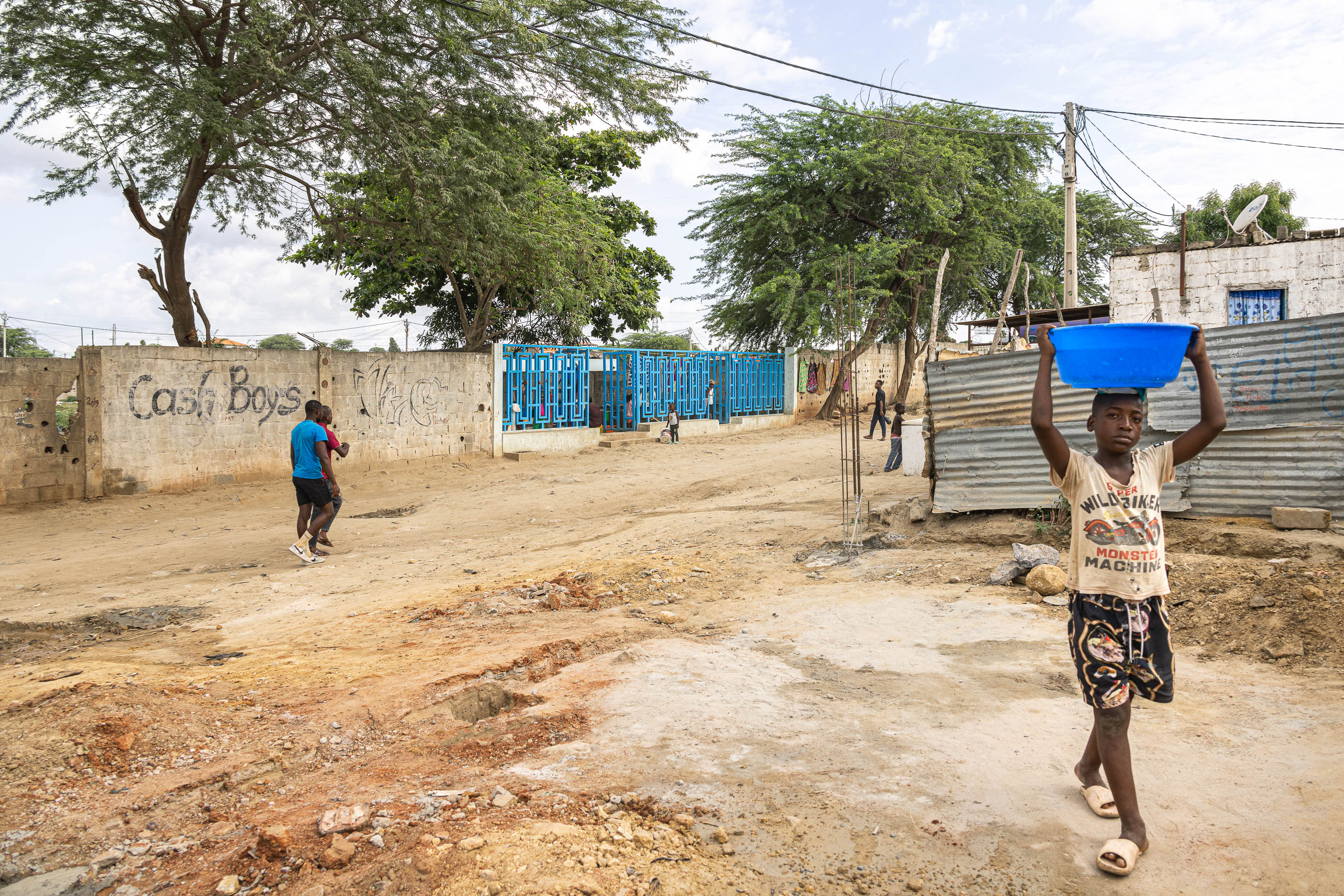 Rua: Rua de terra batida com um chafariz ao fundo, muro de blocos de cimento e casas de zinco. — Chafarizes em Luanda arquitetura Paulo Moreira Architectures em Luanda