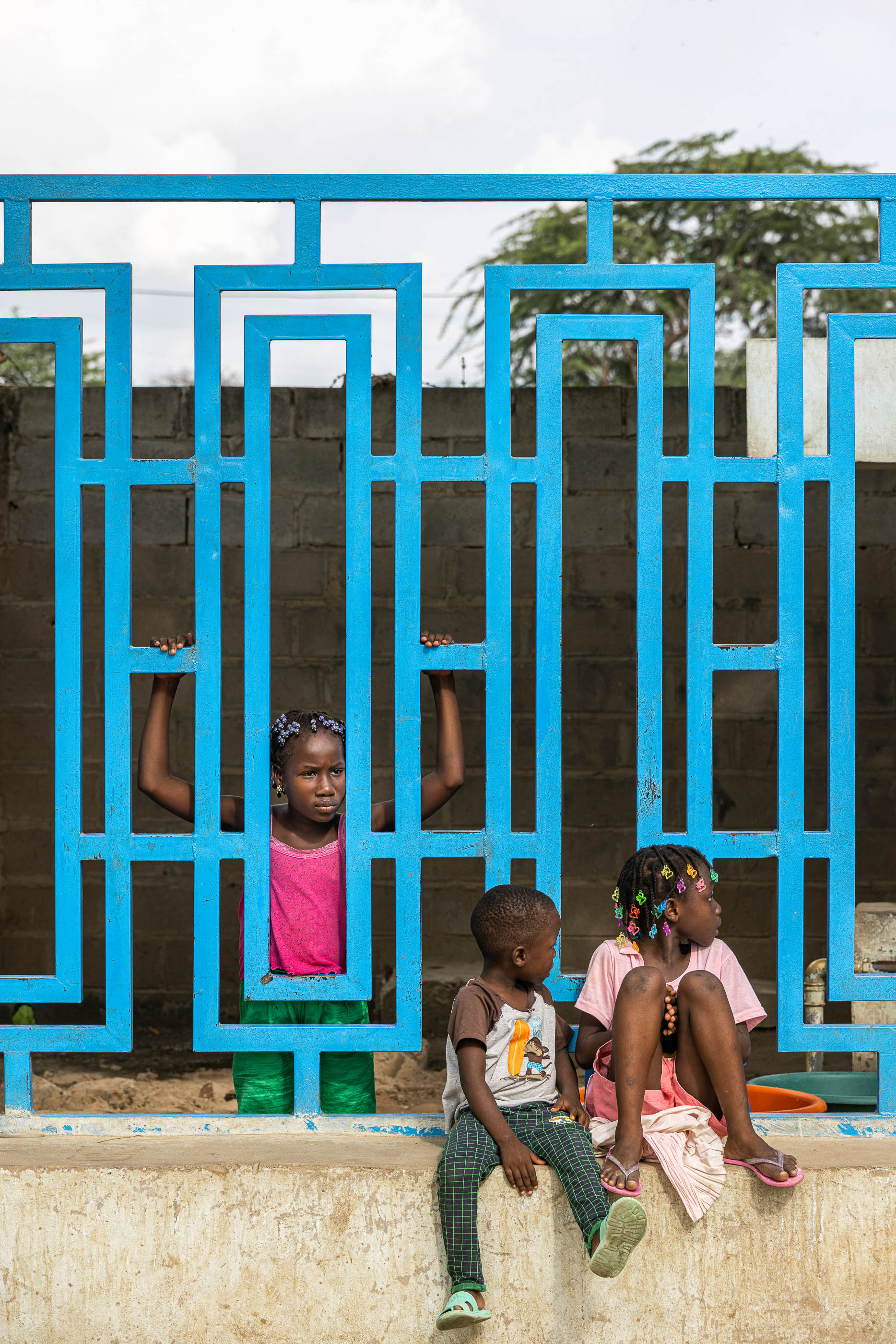 Exterior: Crianças sentadas junto a um chafariz, com grade azul e muro de blocos de cimento. — Chafarizes em Luanda arquitetura Paulo Moreira Architectures em Luanda