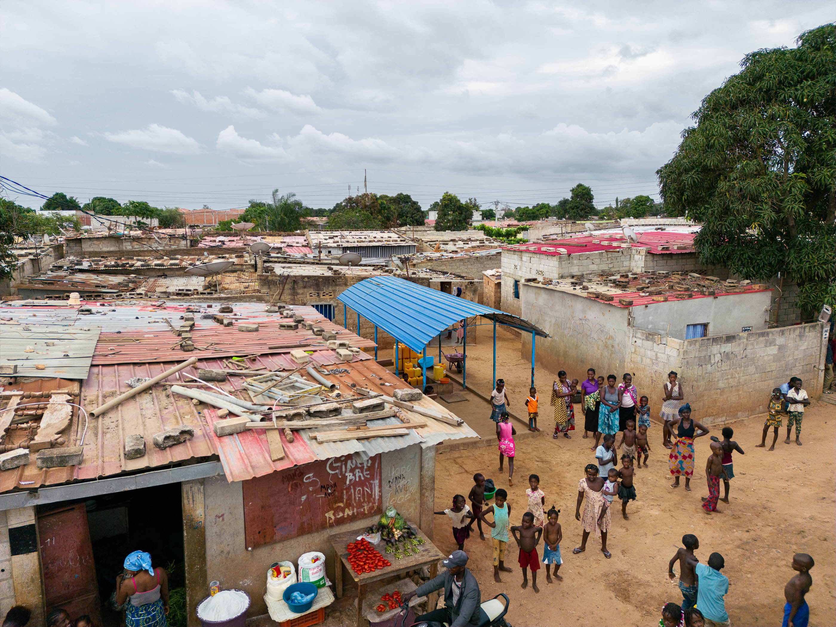 Exterior: Pessoas reunidas junto a um chafariz coberto, com bancas de venda e casas de tijolo à volta. — Chafarizes em Luanda arquitetura Paulo Moreira Architectures em Luanda