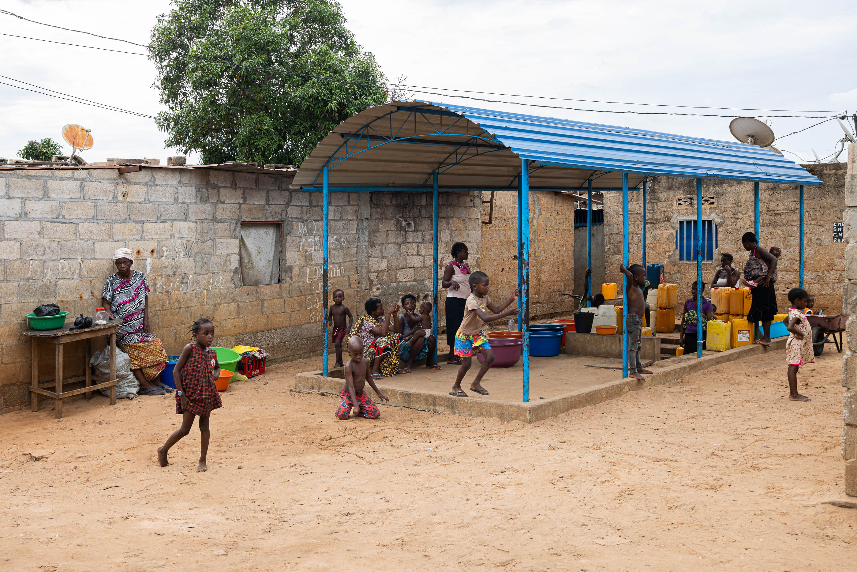 Exterior: Chafariz coberto com pessoas a recolher água, muro de blocos de cimento e bancas de venda. — Chafarizes em Luanda arquitetura Paulo Moreira Architectures em Luanda