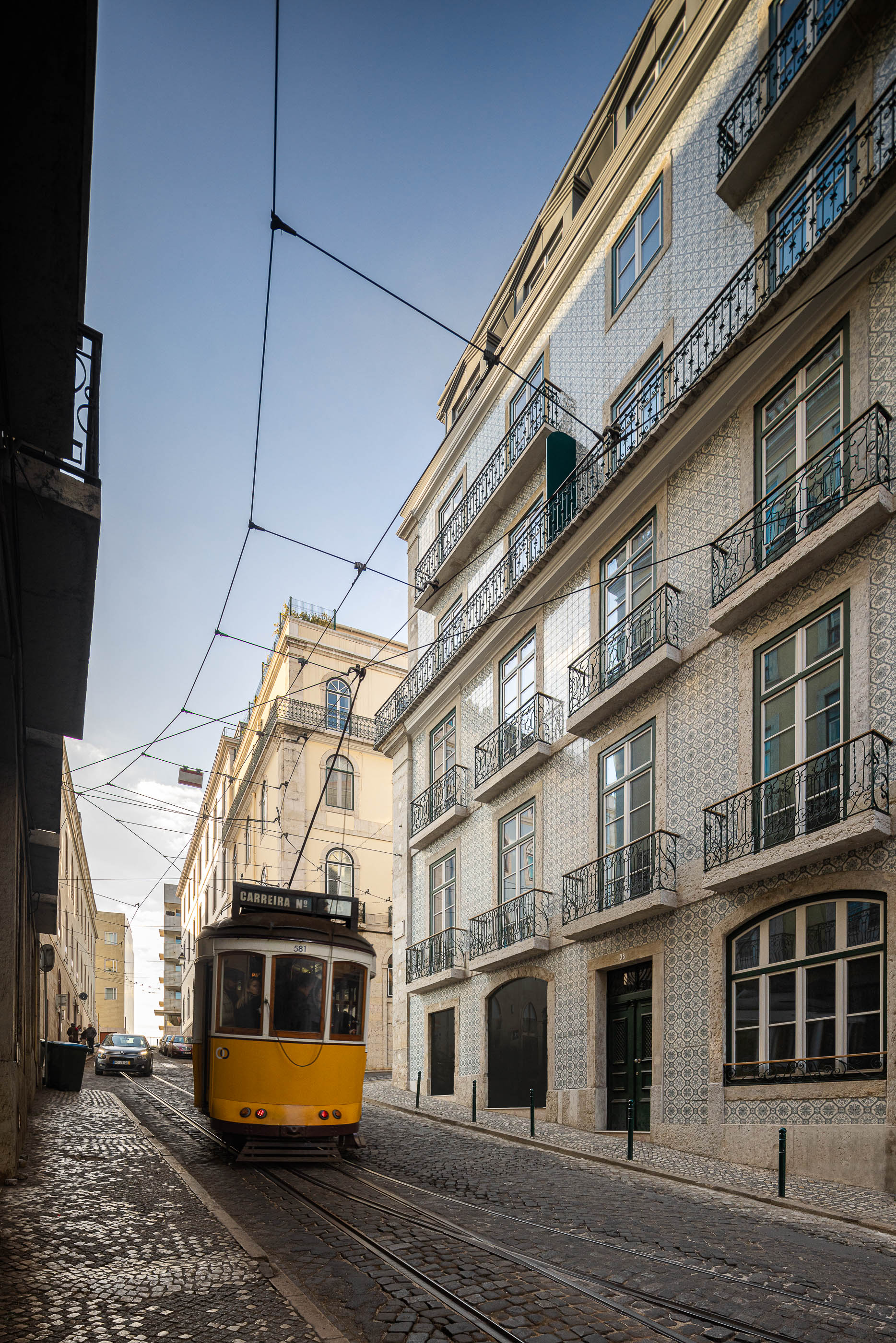 Rua: Elétrico amarelo a passar numa rua calcetada ladeada por edifícios históricos com fachadas em azulejo. — Victor Cordon arquitetura Fragmentos em Lisboa