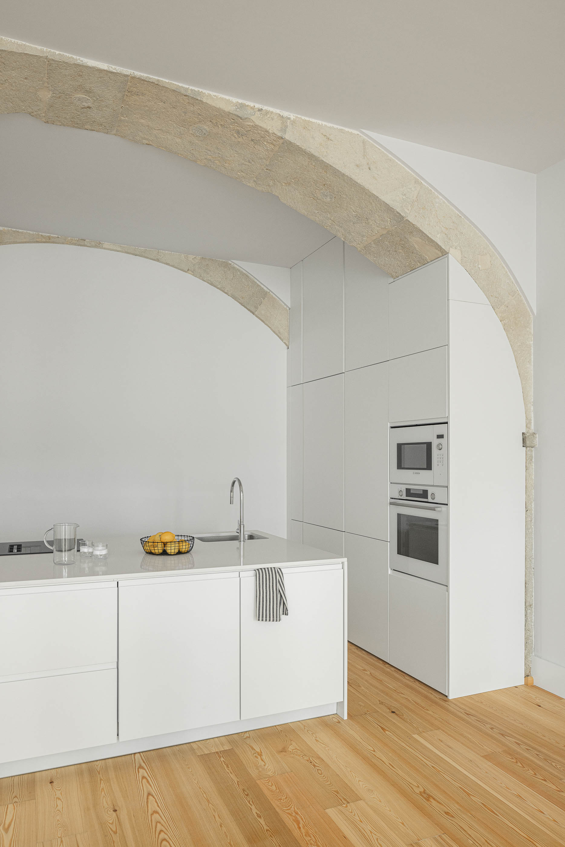 Cozinha: Cozinha moderna com armários brancos minimalistas, bancada em pedra clara e arcos de pedra originais. — Victor Cordon arquitetura Fragmentos em Lisboa