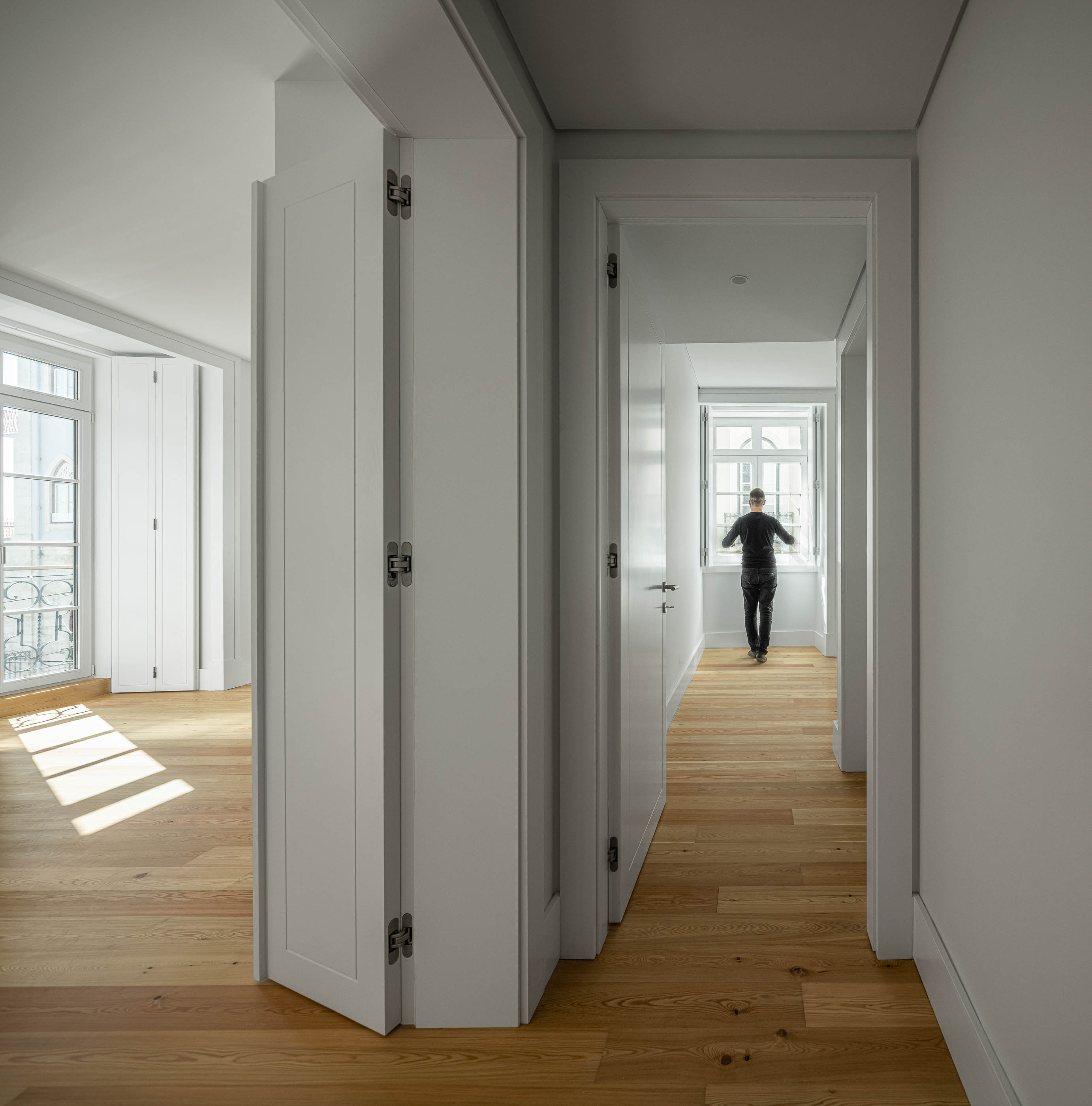 Corredor: Corredor com pavimento em madeira clara, paredes brancas e vista para um quarto com varanda. — Victor Cordon arquitetura Fragmentos em Lisboa