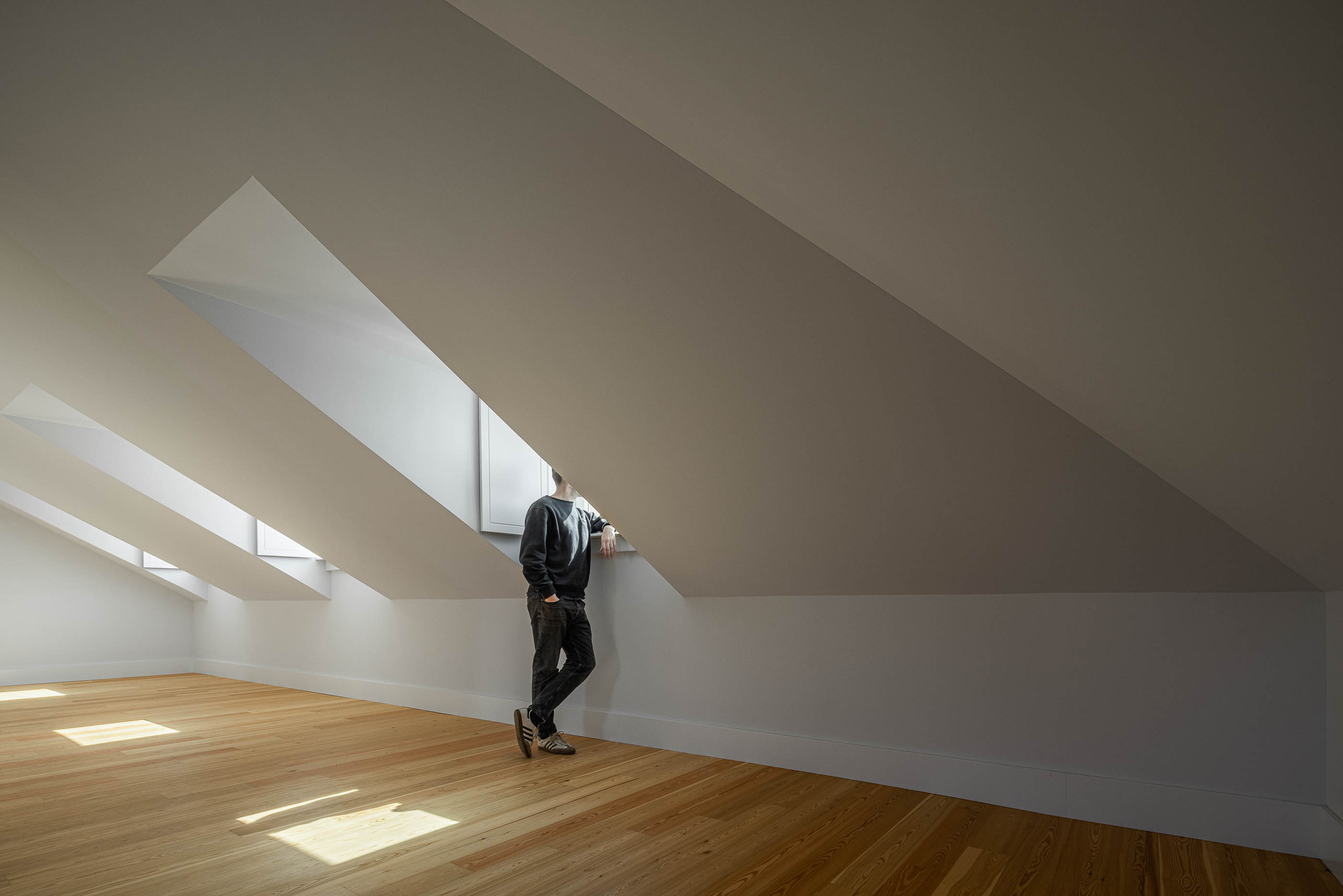 Quarto: Quarto no sótão com pavimento em madeira clara, paredes brancas e janelas triangulares no teto. — Victor Cordon arquitetura Fragmentos em Lisboa