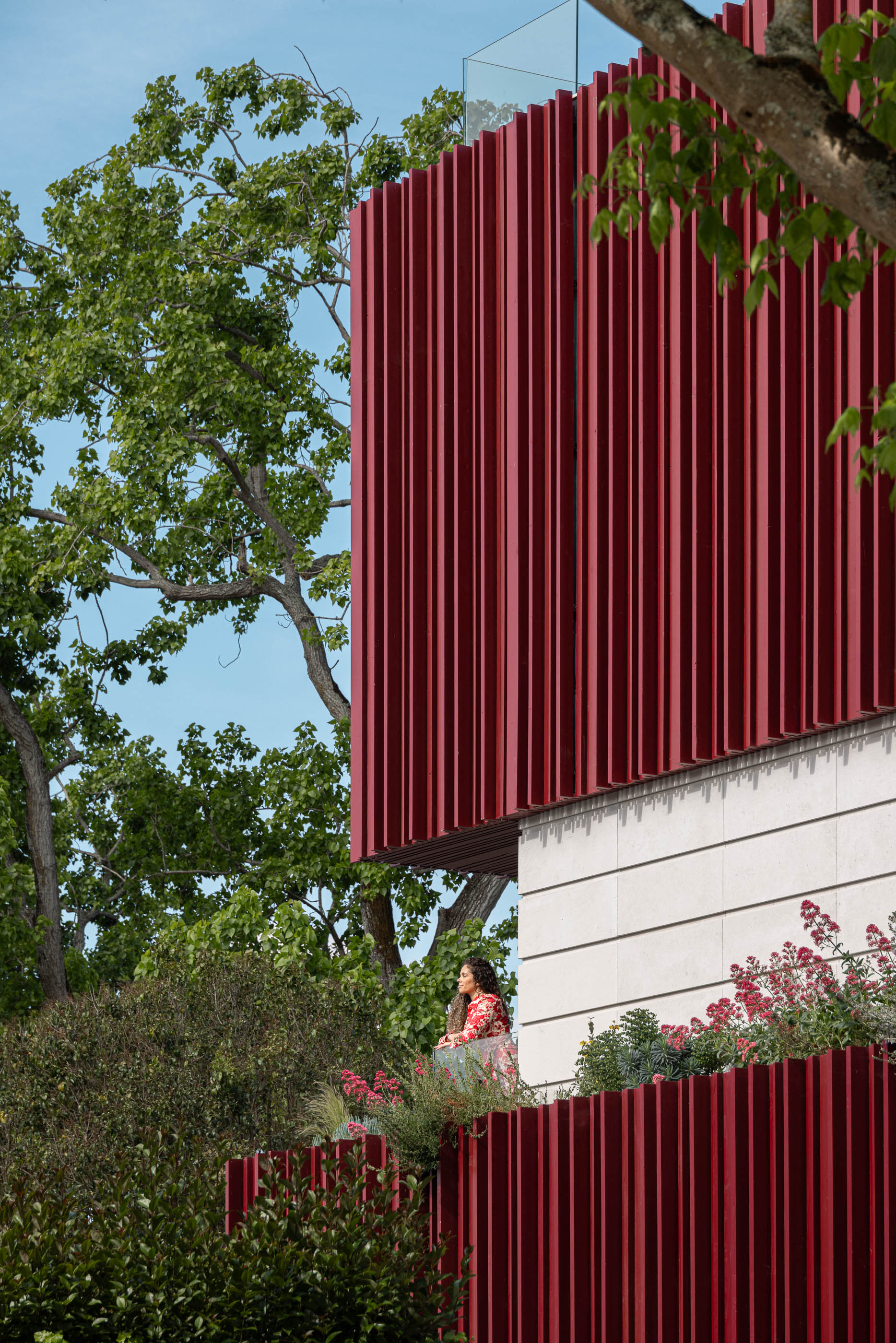 Fachada: Detalhe do revestimento vertical vermelho, varanda com vegetação e mulher a apreciar a vista. — Casa do Parque arquitetura Paulo Martins Arquitectura e Design em Aveiro