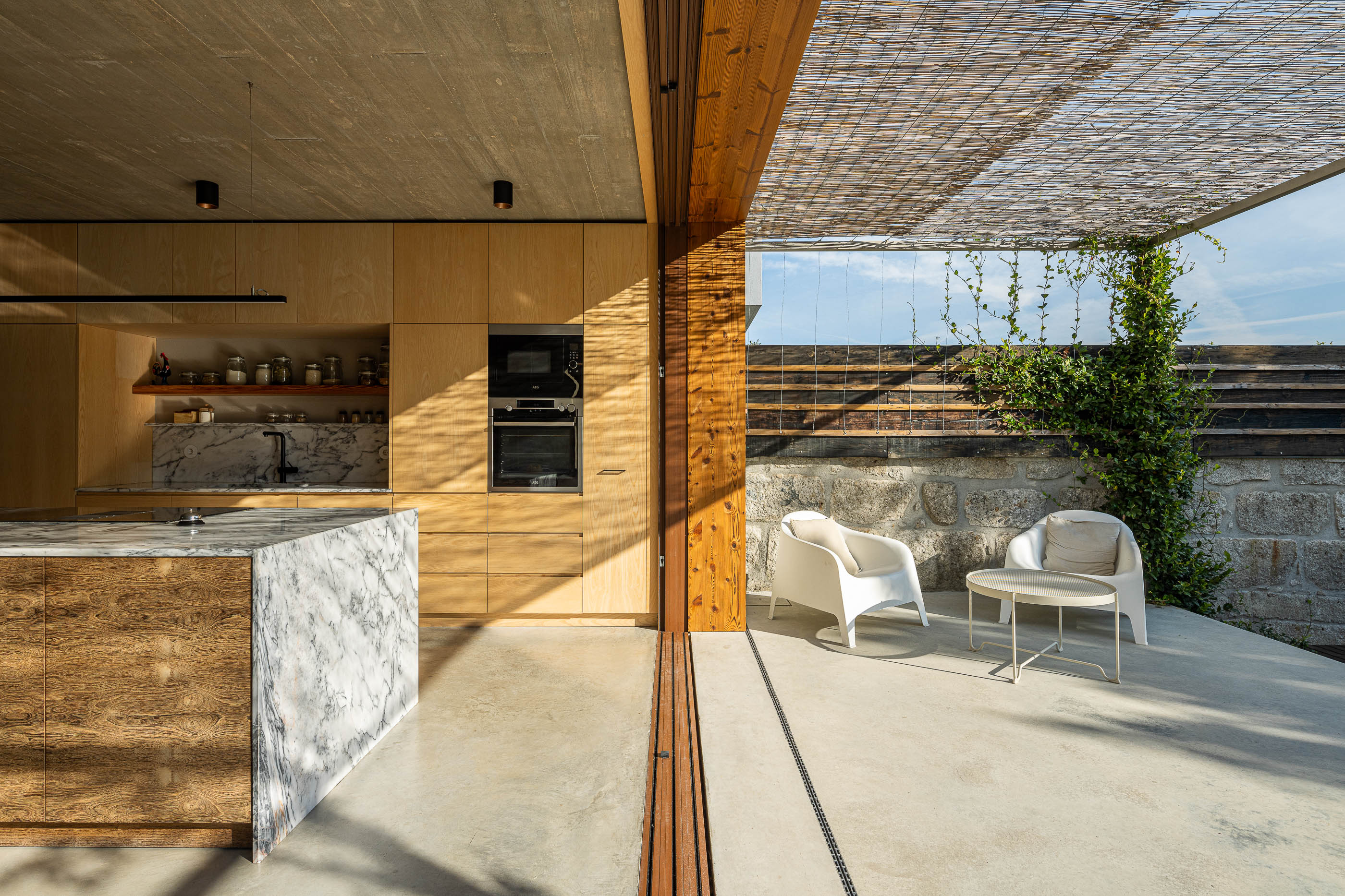 Cozinha/Exterior: Cozinha com acesso direto ao terraço, mobiliário em madeira clara e ilha em mármore. — Casa Fonte Velha arquitetura Martins Pimenta – Arquitetura e Construção em Matosinhos
