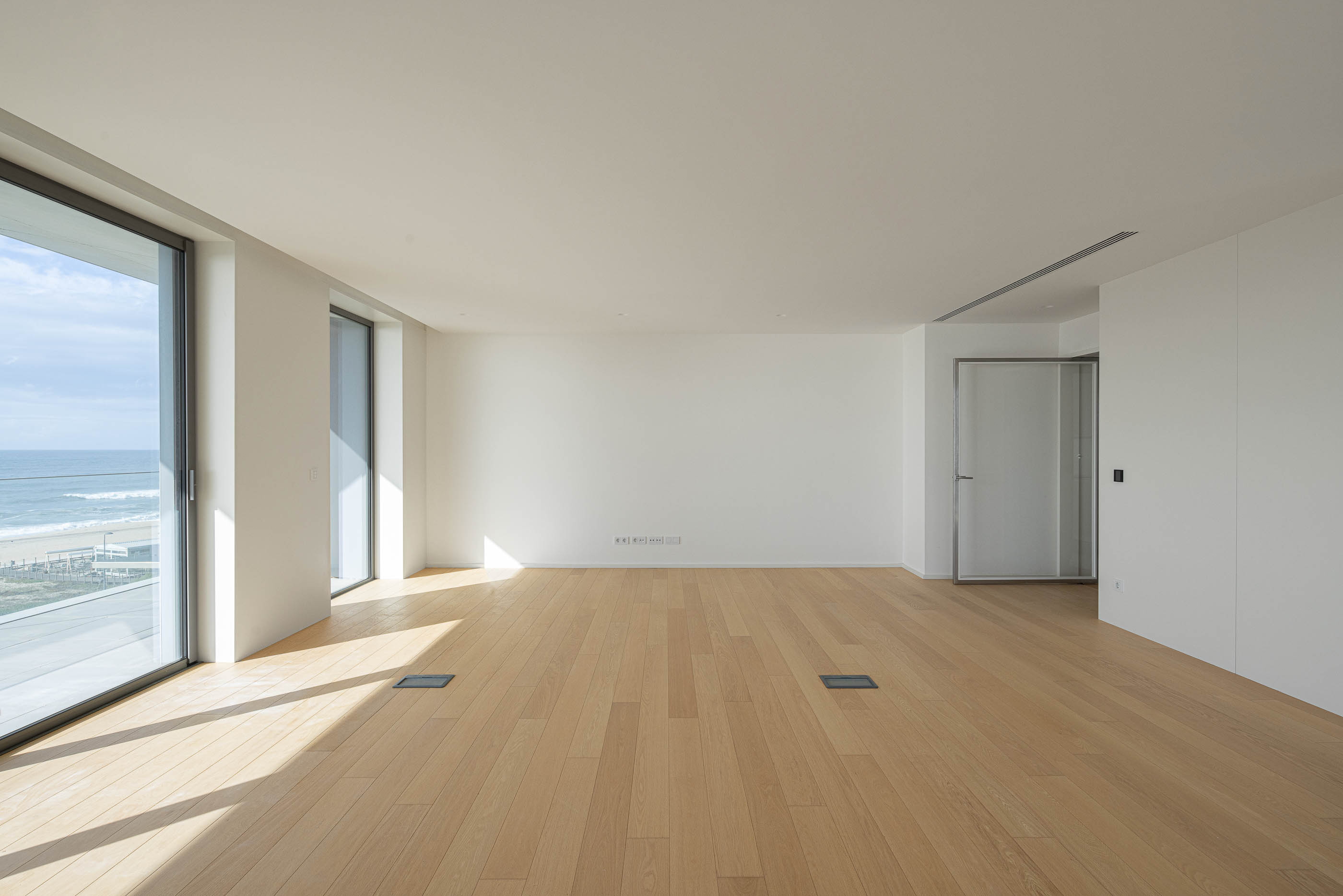Sala de estar: Espaço amplo e luminoso com pavimento em madeira e paredes brancas. — Edifício Seashore arquitetura Atelier d´Arquitectura Lopes da Costa em Vila Nova de Gaia