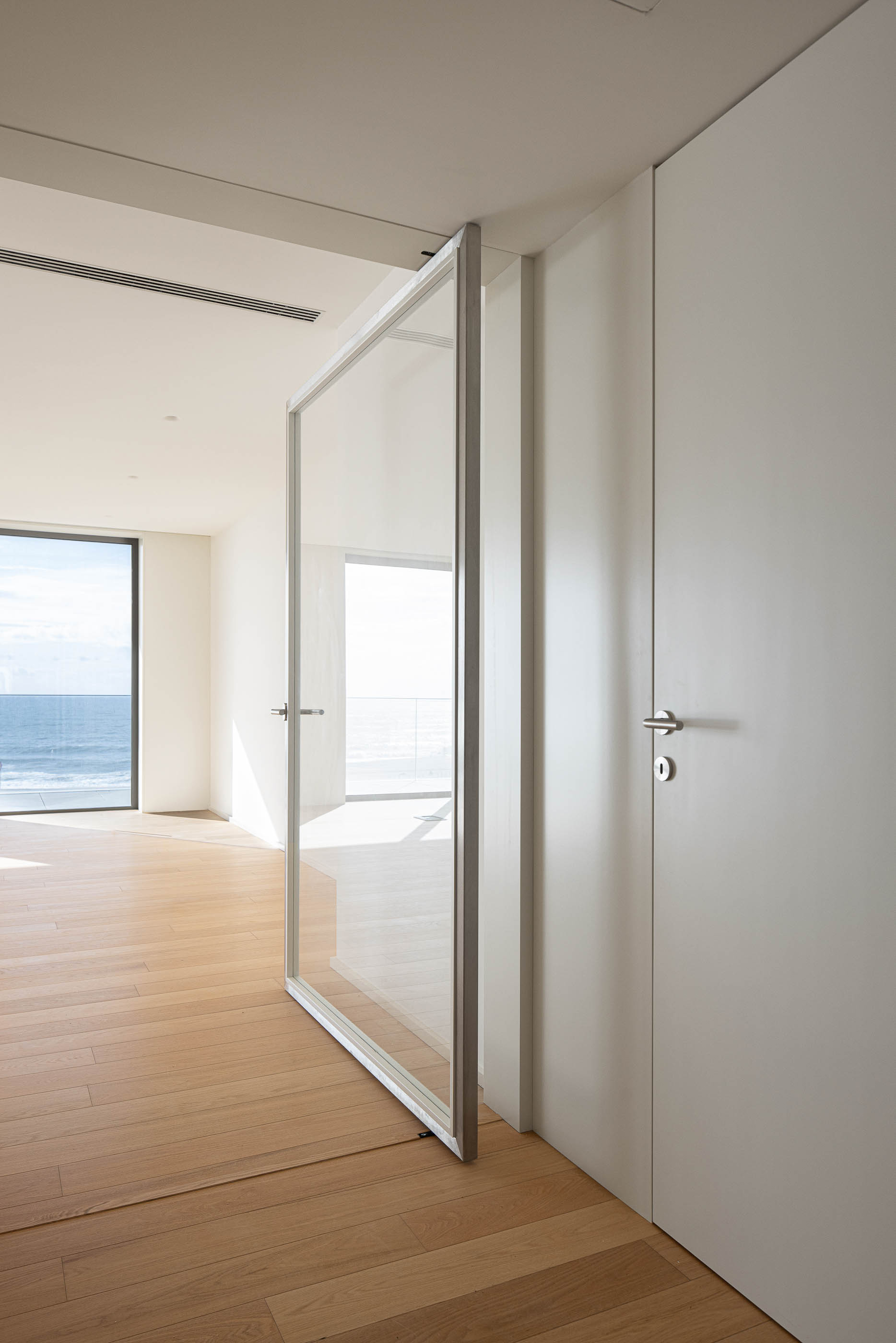 Sala de estar: Porta pivotante com estrutura metálica e pavimento em madeira. — Edifício Seashore arquitetura Atelier d´Arquitectura Lopes da Costa em Vila Nova de Gaia