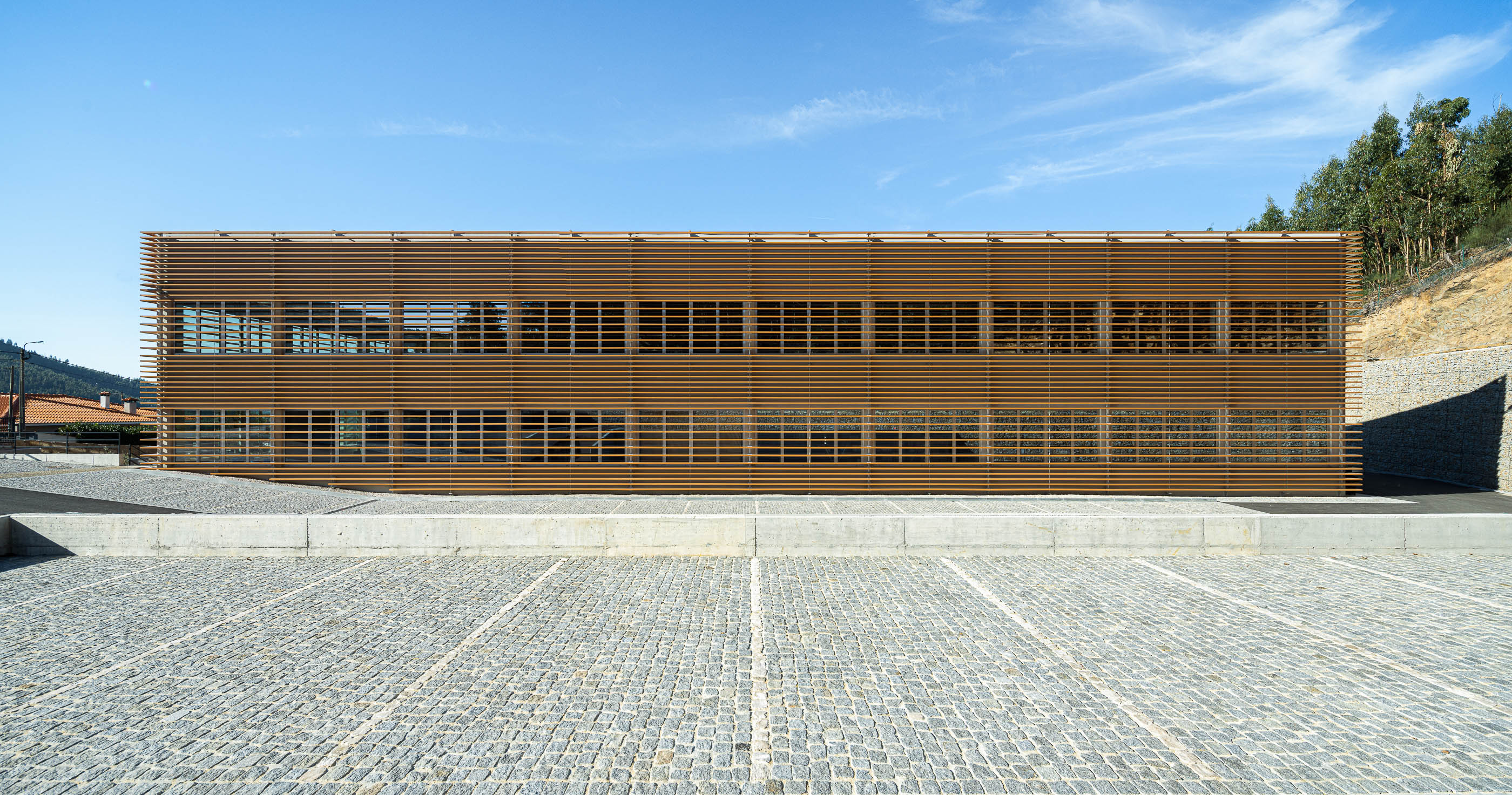 Fachada: Edifício industrial com fachada em ripas de madeira, estacionamento em calçada e muro de suporte em betão. — Unidade Industrial OROPOL arquitetura Em Paralelo em Castelo de Paiva