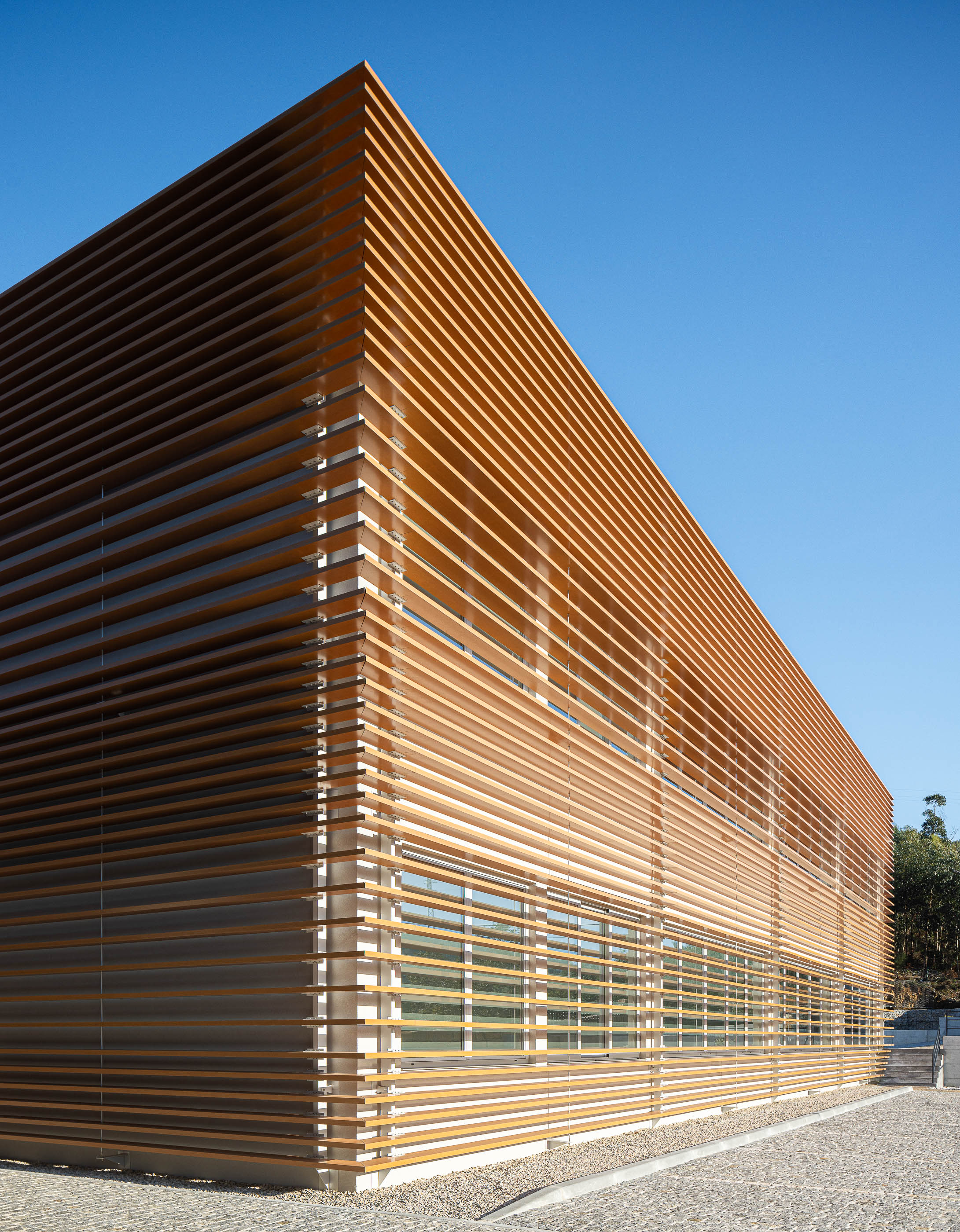 Fachada: Detalhe da esquina do edifício industrial com revestimento em ripas de madeira e pavimento em calçada. — Unidade Industrial OROPOL arquitetura Em Paralelo em Castelo de Paiva