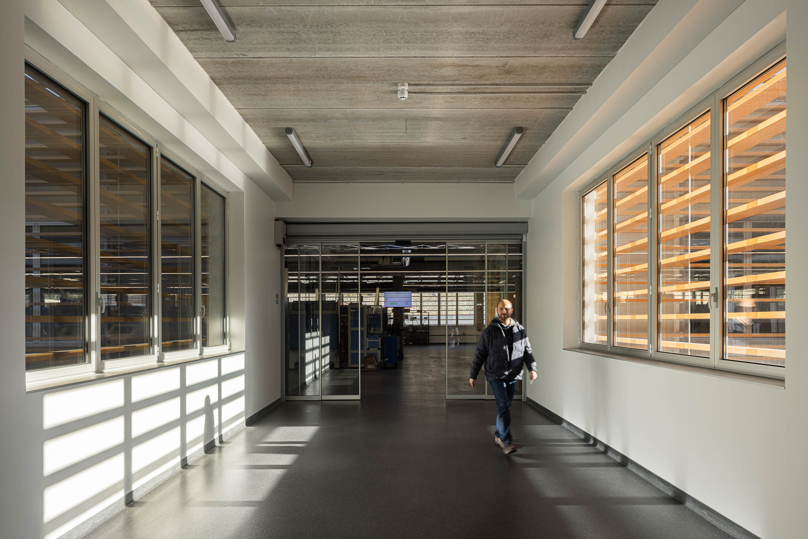 Corredor: Espaço de circulação com pavimento em borracha, paredes brancas, janelas com ripas de madeira e porta de vidro. — Unidade Industrial OROPOL arquitetura Em Paralelo em Castelo de Paiva