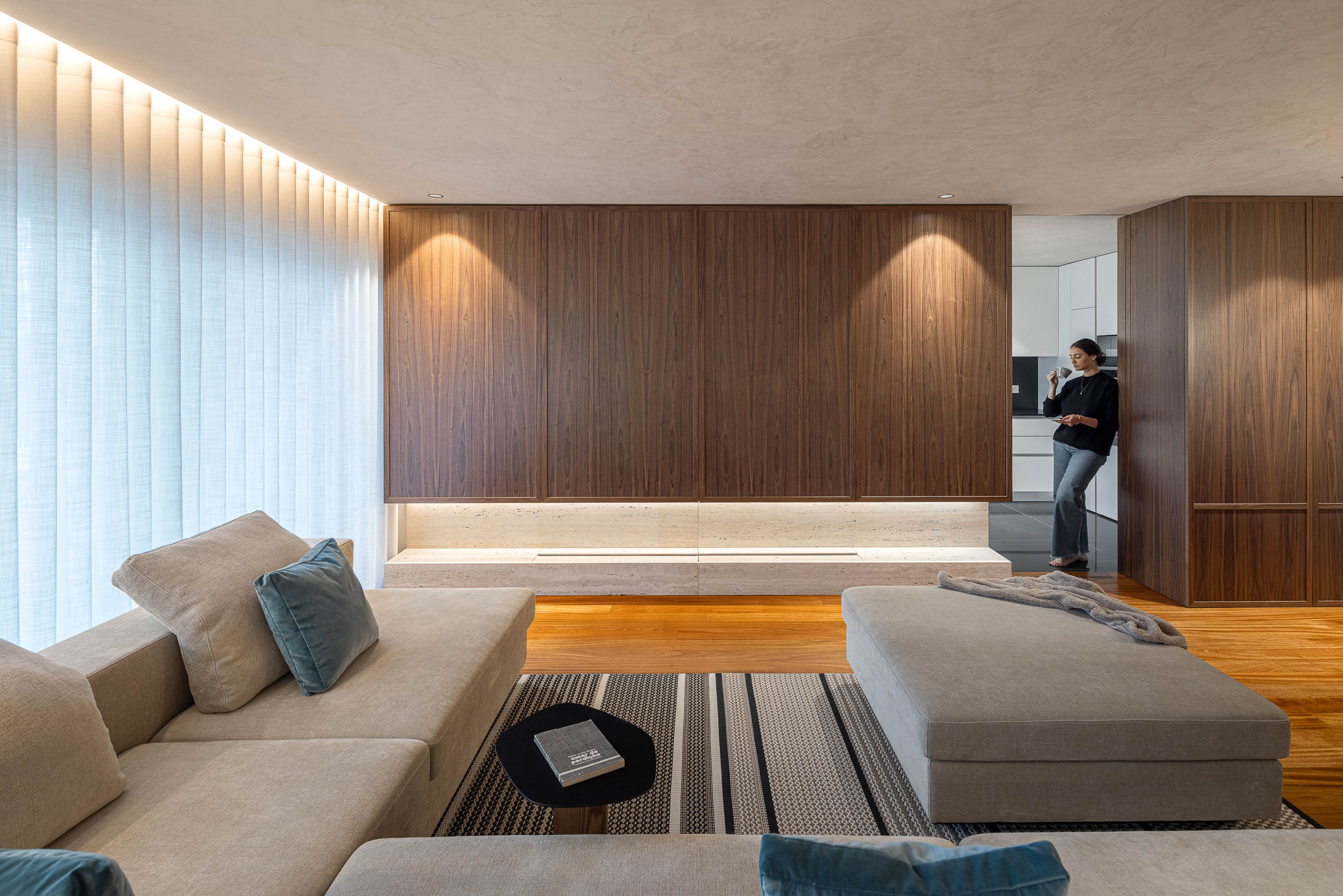 Sala de estar: Sofá modular bege com almofadas azuis, parede revestida a madeira e nicho em travertino iluminado. — Lugar Comum arquitetura edmundo* architecture + planning em vila do conde