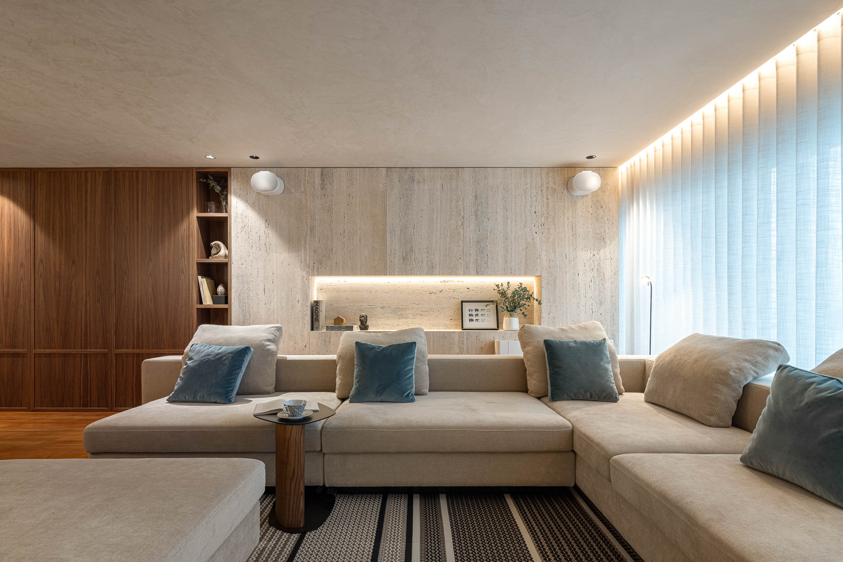 Sala de estar: Sofá modular bege com almofadas azuis, parede em travertino e estantes em madeira. — Lugar Comum arquitetura edmundo* architecture + planning em vila do conde