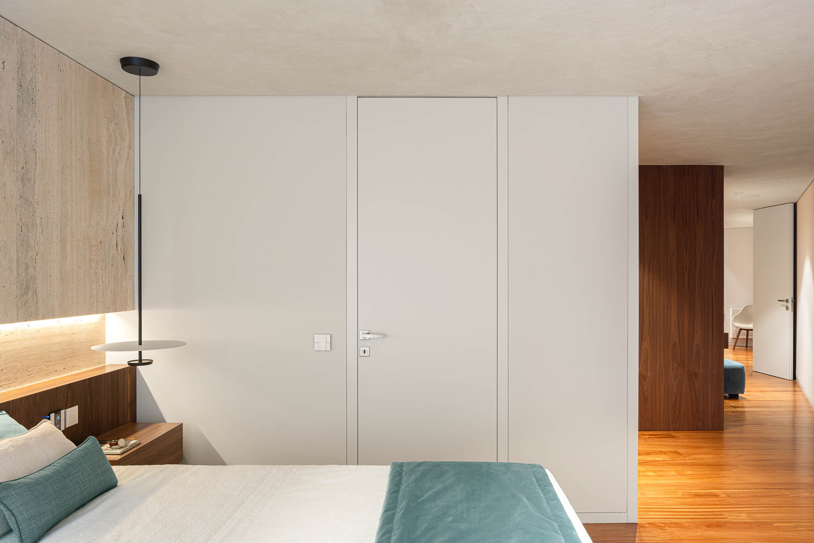 Quarto: Cama com cabeceira em madeira e travertino, candeeiro suspenso e porta branca. — Lugar Comum arquitetura edmundo* architecture + planning em vila do conde