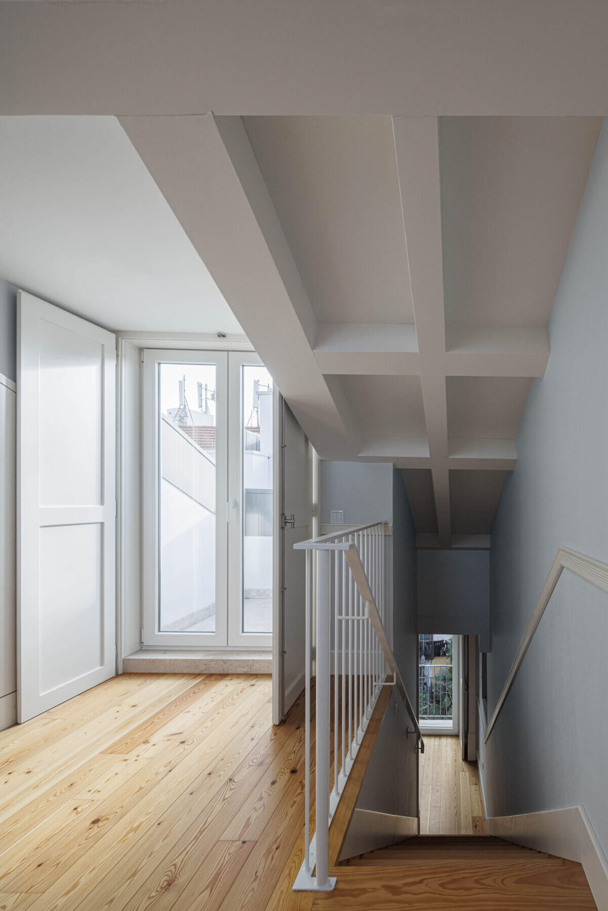 Sótão: Sótão com pavimento em madeira, teto com vigas brancas e acesso a varanda. — Villa Chiado arquitetura Pedro Carrilho Arquitectos em Lisboa