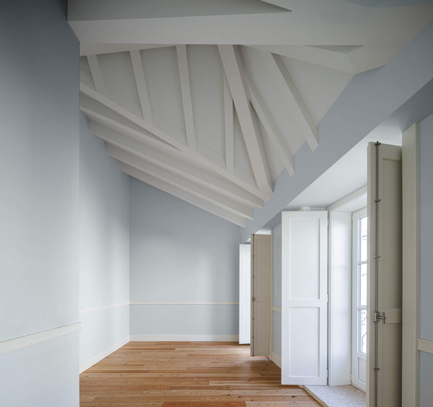 Sótão: Sótão com paredes azul claro, pavimento em madeira e teto com vigas brancas. — Villa Chiado arquitetura Pedro Carrilho Arquitectos em Lisboa
