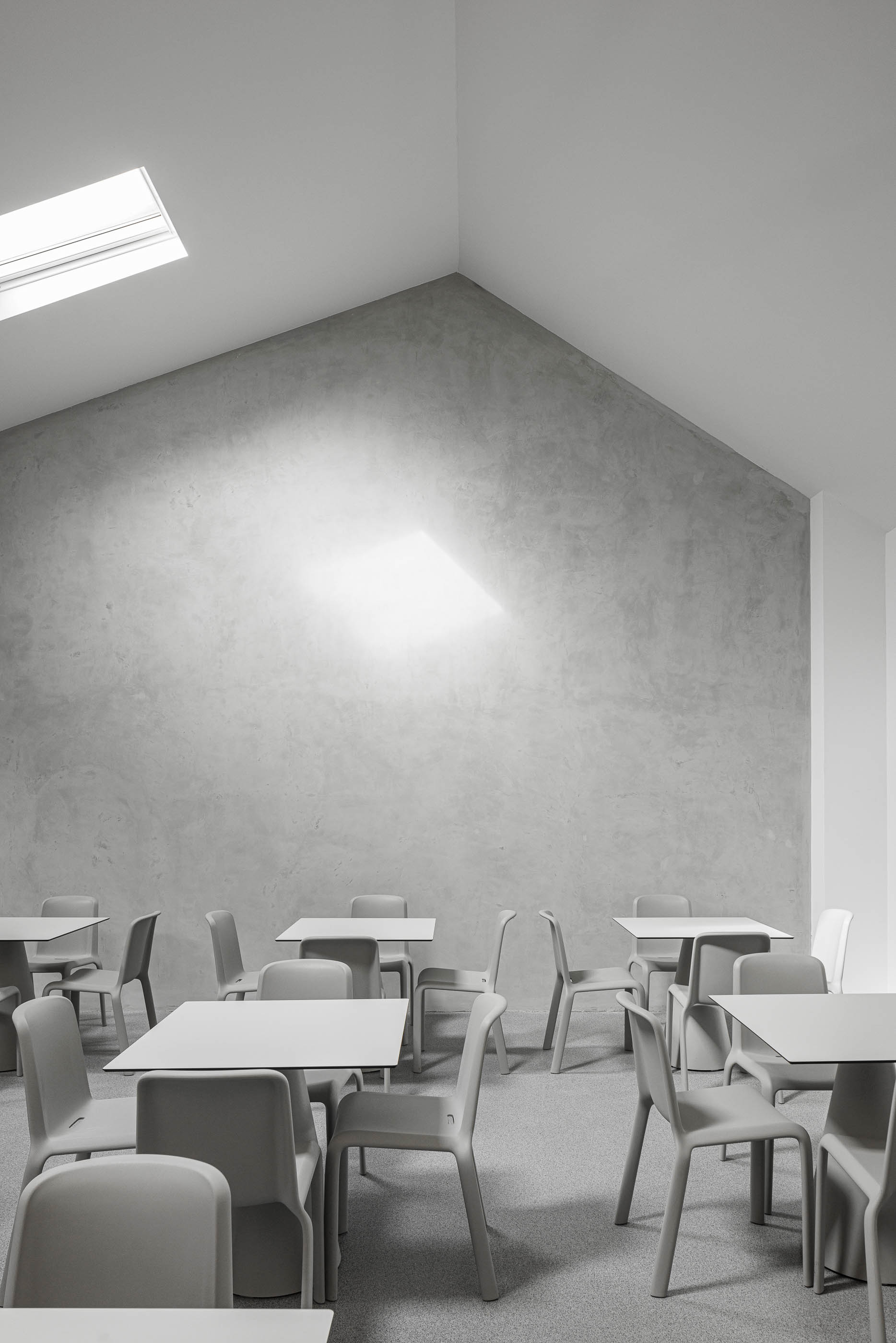 Refeitório: Paredes em cimento afagado sob telhado inclinado com claraboia, mesas e cadeiras cinzentas sobre pavimento cinzento. — Indústria | A10 arquitetura BOX arquitectos em São Miguel