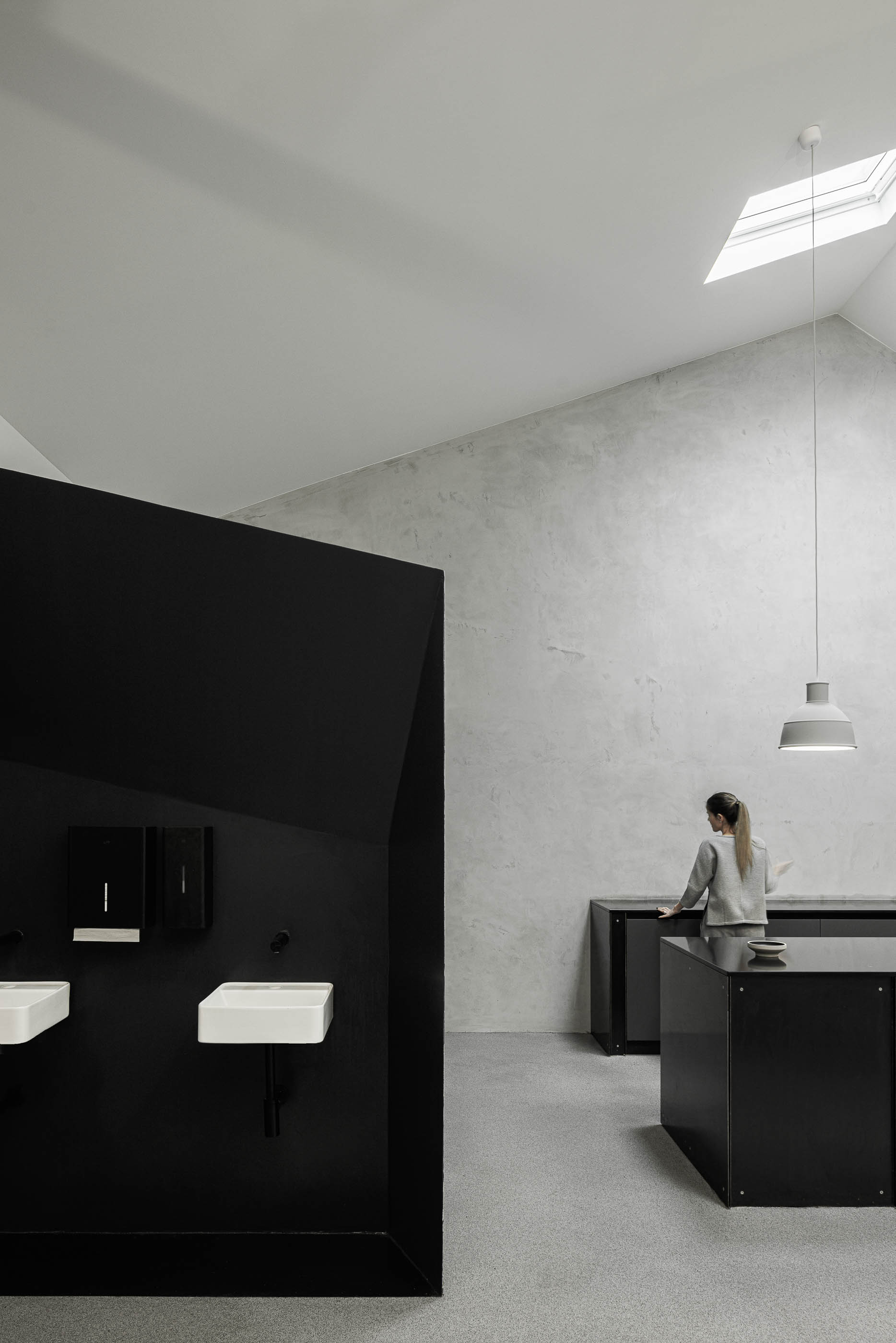 Casa de banho: Lavatórios brancos em parede preta geométrica, com balcão preto e candeeiro pendurado, pavimento cinzento. — Indústria | A10 arquitetura BOX arquitectos em São Miguel