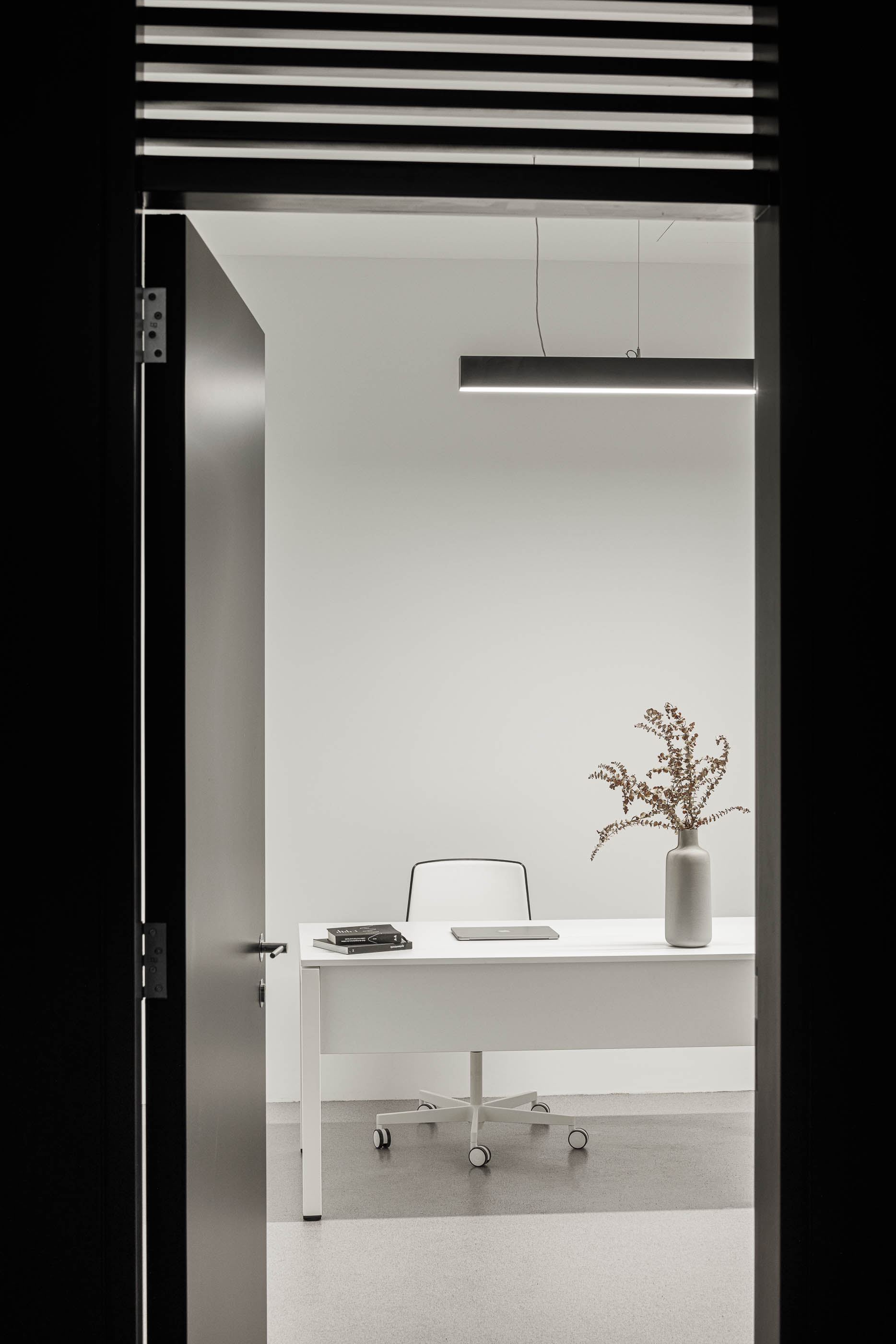 Escritório: Vista através de porta preta para secretária branca com candeeiro suspenso e vaso com flores. — Indústria | A10 arquitetura BOX arquitectos em São Miguel