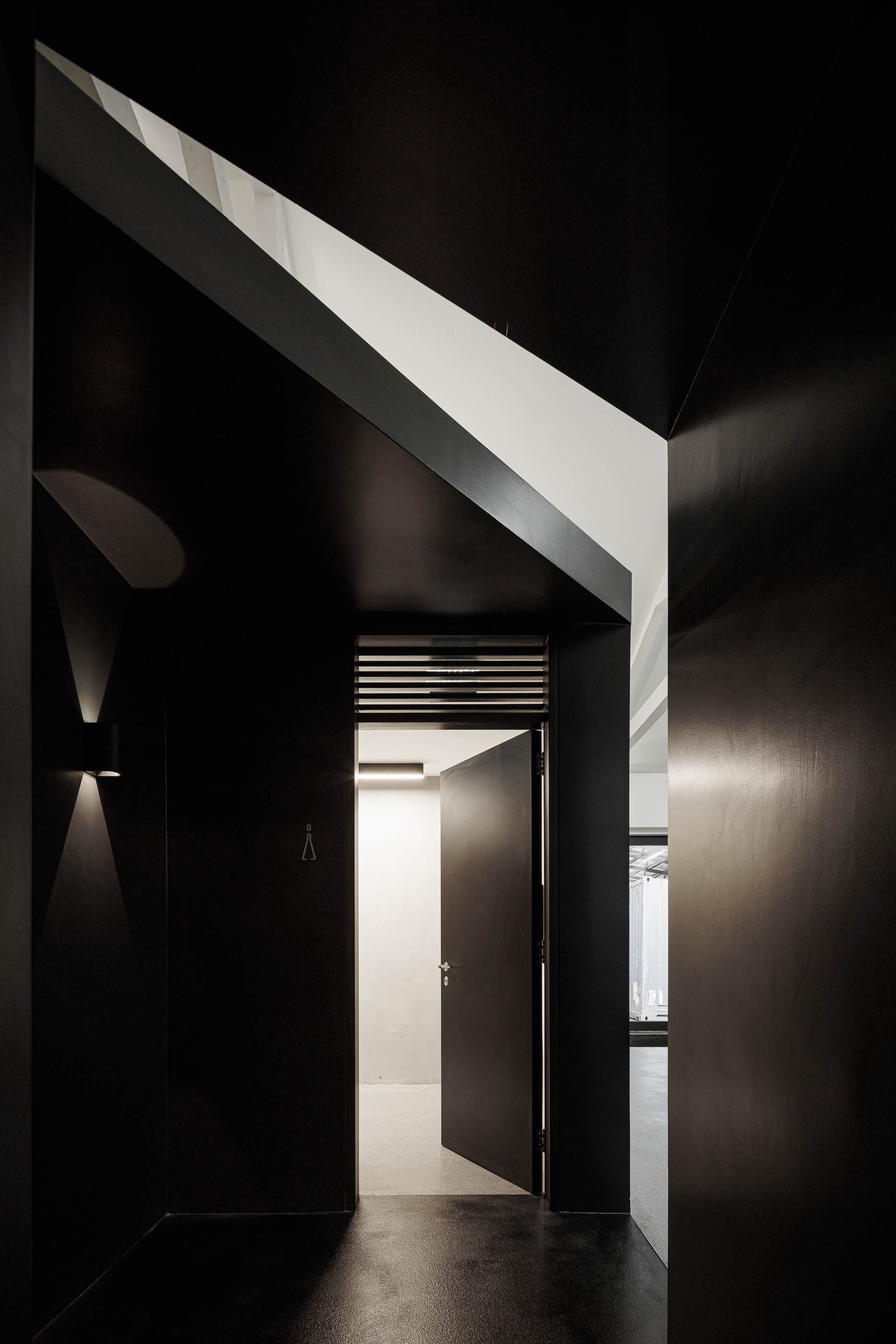 Corredor: Porta preta aberta com candeeiro de parede, pavimento preto e vista para corredor com paredes em cimento afagado. — Indústria | A10 arquitetura BOX arquitectos em São Miguel
