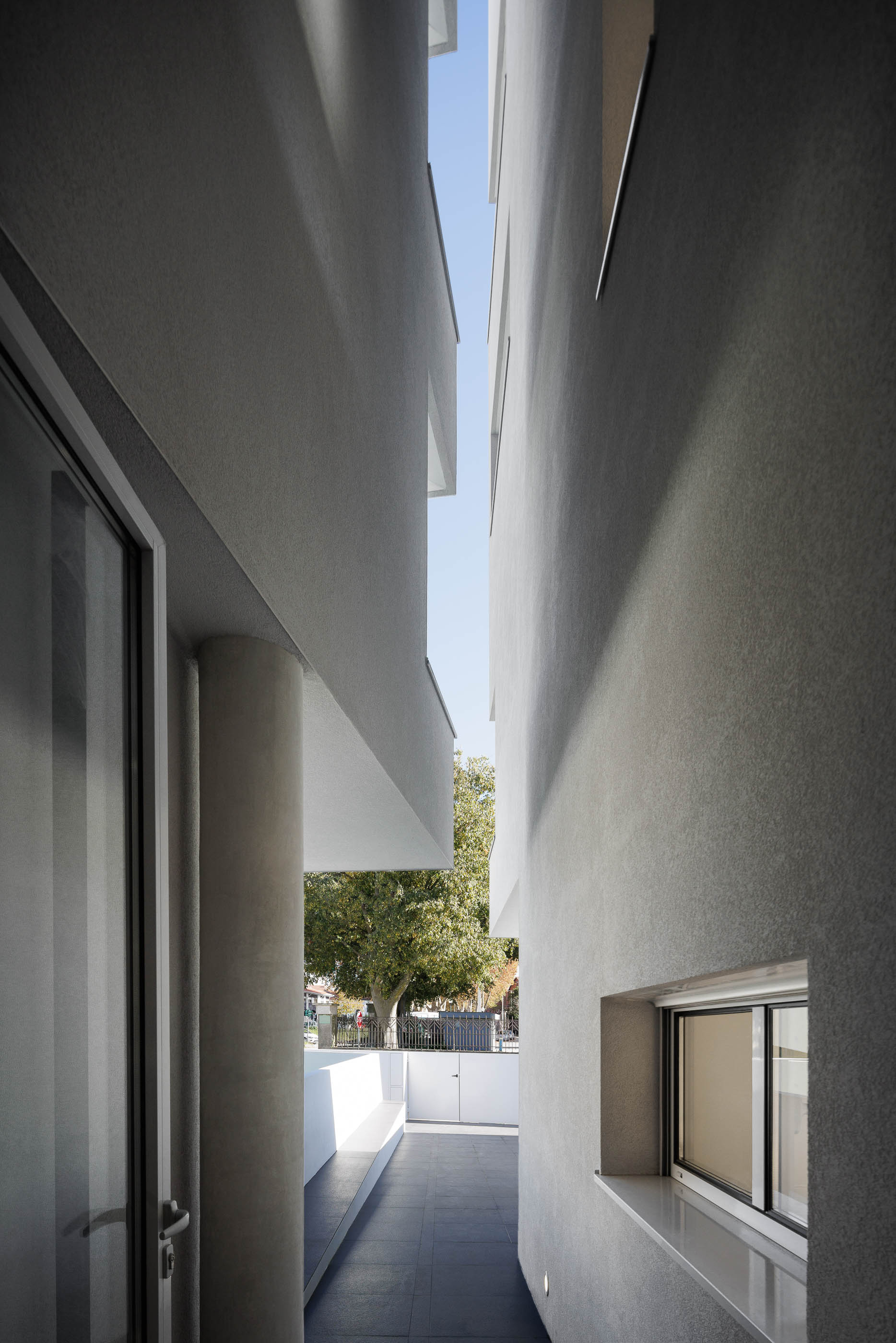 Corredor exterior: Espaço estreito entre dois edifícios brancos, com pavimento escuro e vista para a rua. — Nova Rio Habitação Multifamiliar arquitetura António Paulo Marques, Arquiteto e Associados em Porto