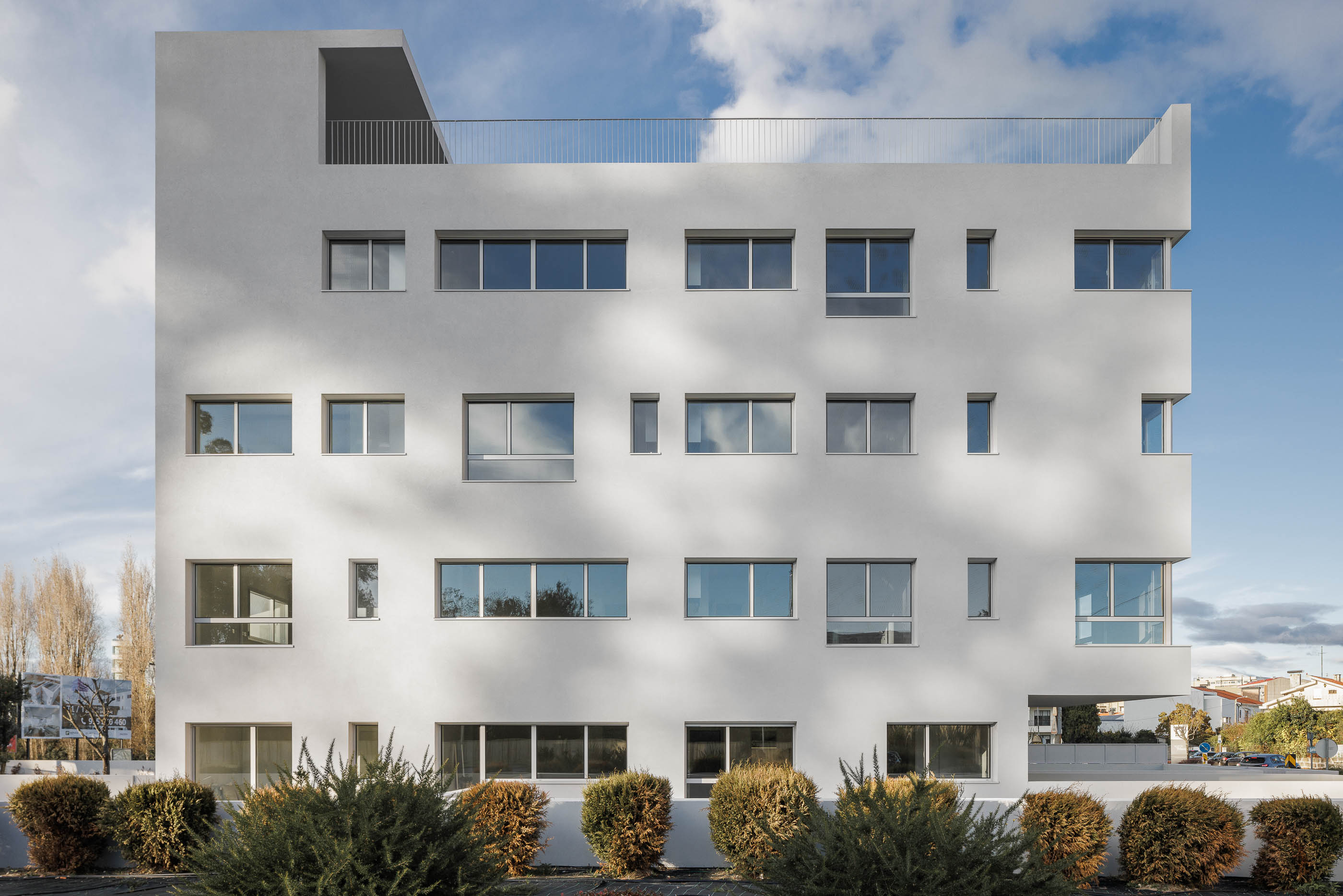 Fachada: Edifício branco com janelas retangulares, terraço no topo e vegetação em primeiro plano. — Nova Rio Habitação Multifamiliar arquitetura António Paulo Marques, Arquiteto e Associados em Porto