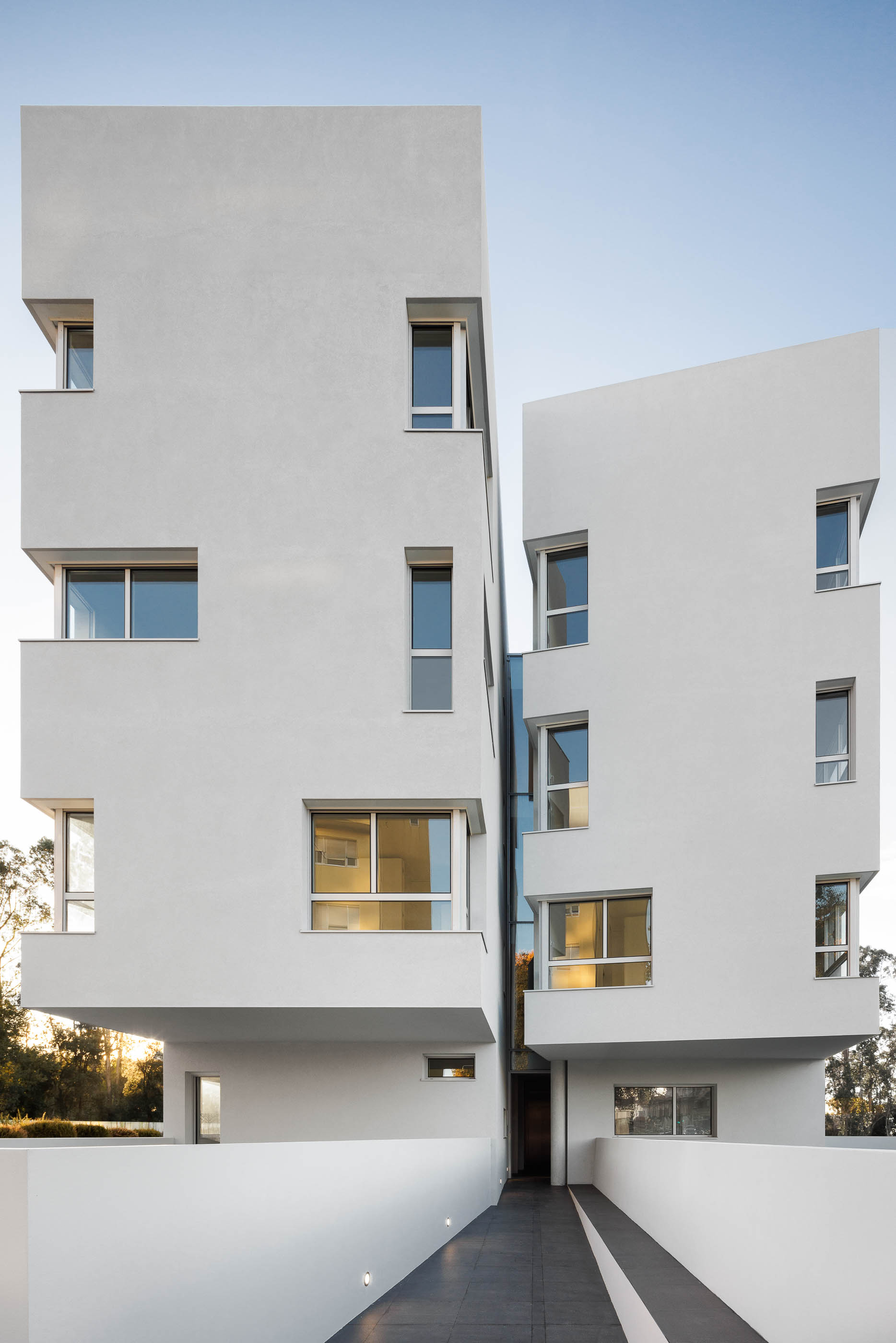 Fachada: Edifício branco com janelas retangulares e passagem envidraçada, com vegetação ao fundo. — Nova Rio Habitação Multifamiliar arquitetura António Paulo Marques, Arquiteto e Associados em Porto