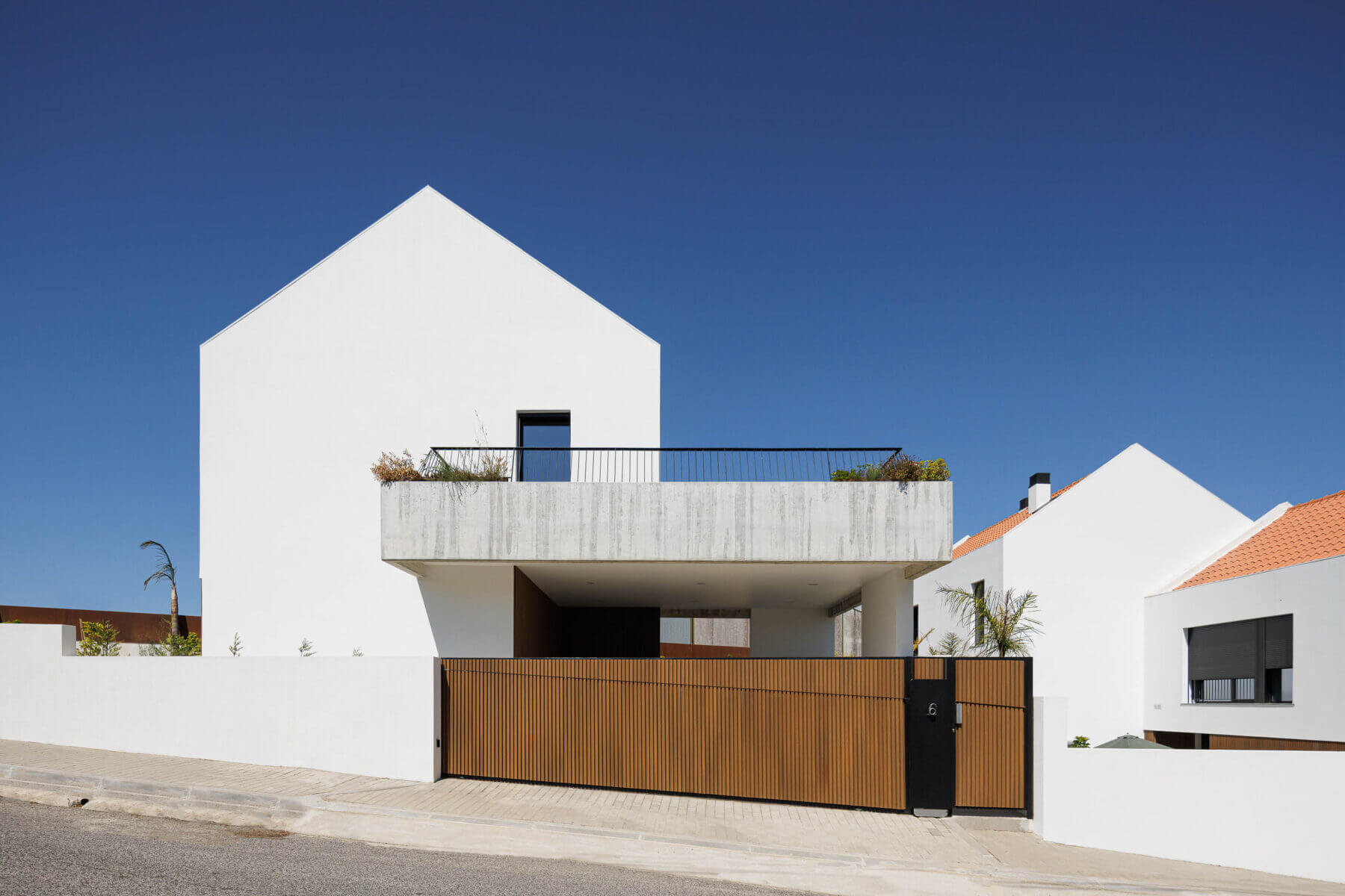 Fachada: Moradia minimalista branca com portão de madeira ripada e varanda em betão aparente com vegetação. — Casas BD arquitetura Esquissos Arquitectura em Sintra