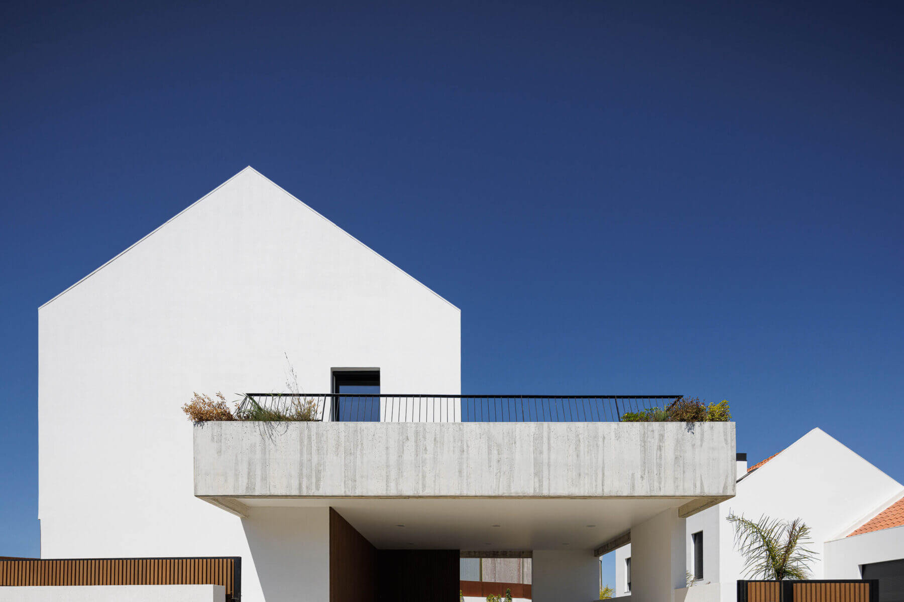 Fachada: Detalhe da varanda em betão aparente com gradeamento preto e vegetação, contra um céu azul. — Casas BD arquitetura Esquissos Arquitectura em Sintra