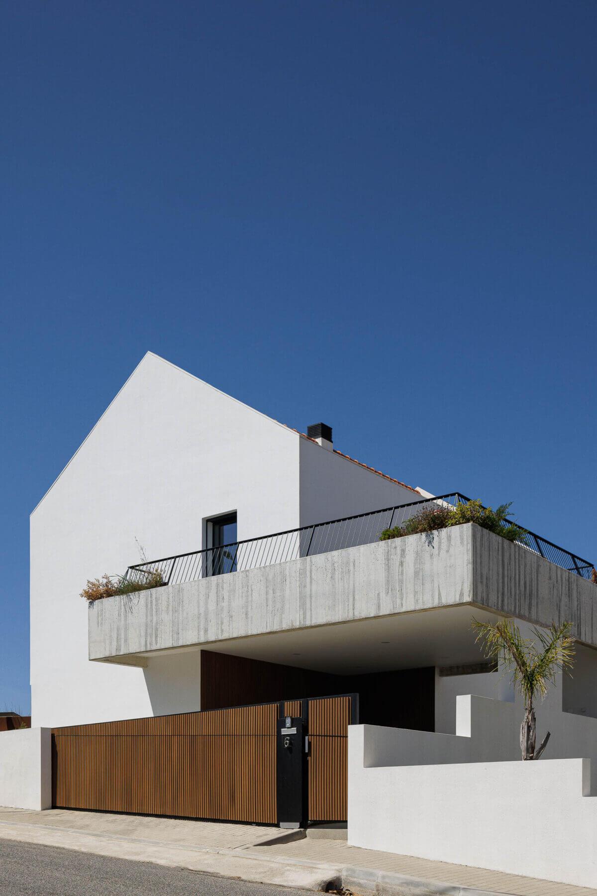 Fachada: Moradia branca com varanda em betão aparente, portão de madeira ripada e jardim frontal. — Casas BD arquitetura Esquissos Arquitectura em Sintra