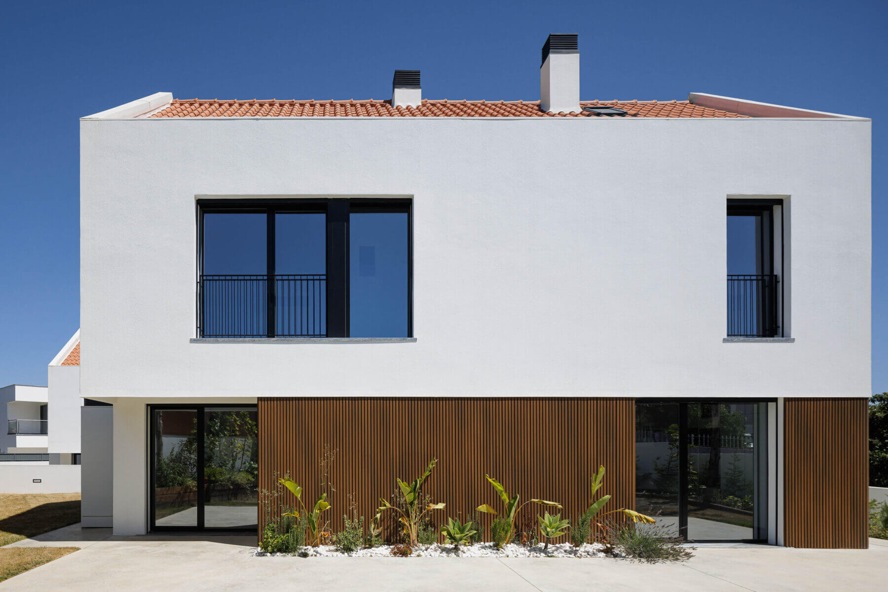 Fachada: Vista lateral da moradia com revestimento em madeira, janelas pretas e jardim com seixos brancos. — Casas BD arquitetura Esquissos Arquitectura em Sintra